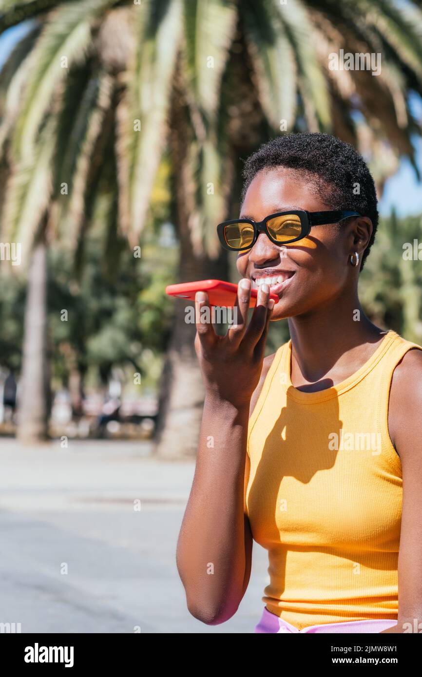 Stilvolle afrikanische Frau, die an einem sonnigen Tag eine Sprachnachricht im Freien sendet. Sie trägt lässige Sommerkleider und lächelt fröhlich Stockfoto