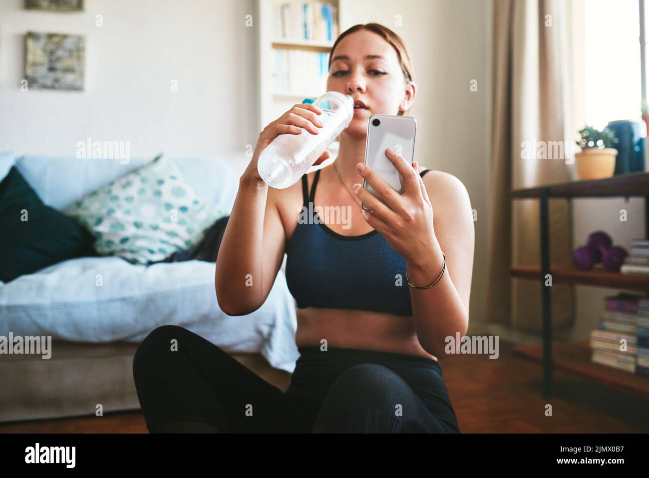 Schnelle Wasser- und Social-Media-Pause. Eine attraktive junge Frau sitzt und trinkt Wasser, während sie ihr Handy nach dem Training benutzt. Stockfoto