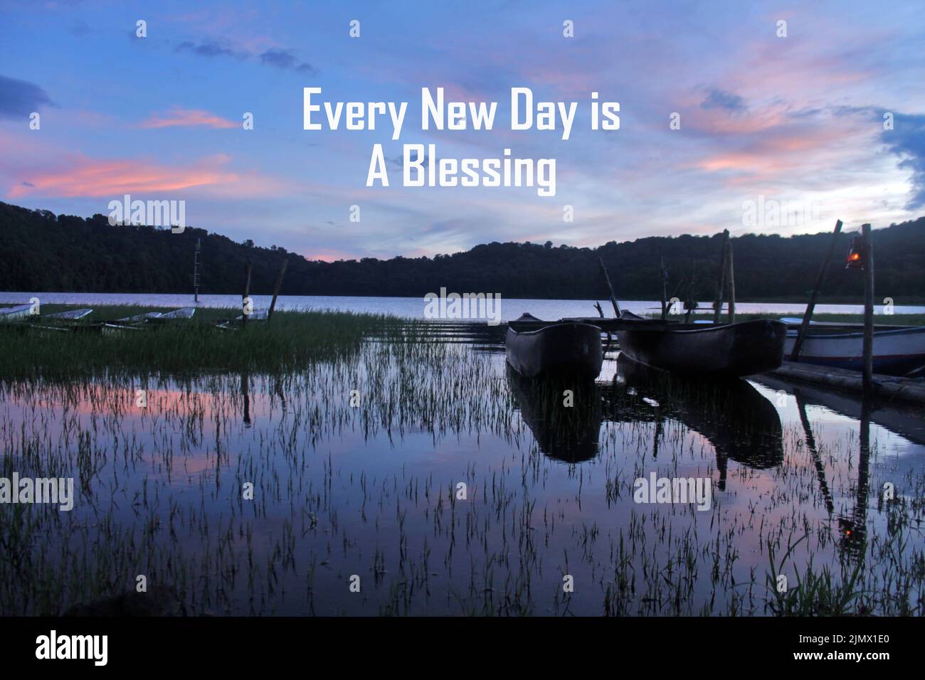 Inspirierendes Morgenzitat - jeder neue Tag ist Ein Segen. Mit blauem Sonnenaufgang über dem See mit hölzernen Fischerbooten, grünem Gras und seiner Wasserspiegelung Stockfoto