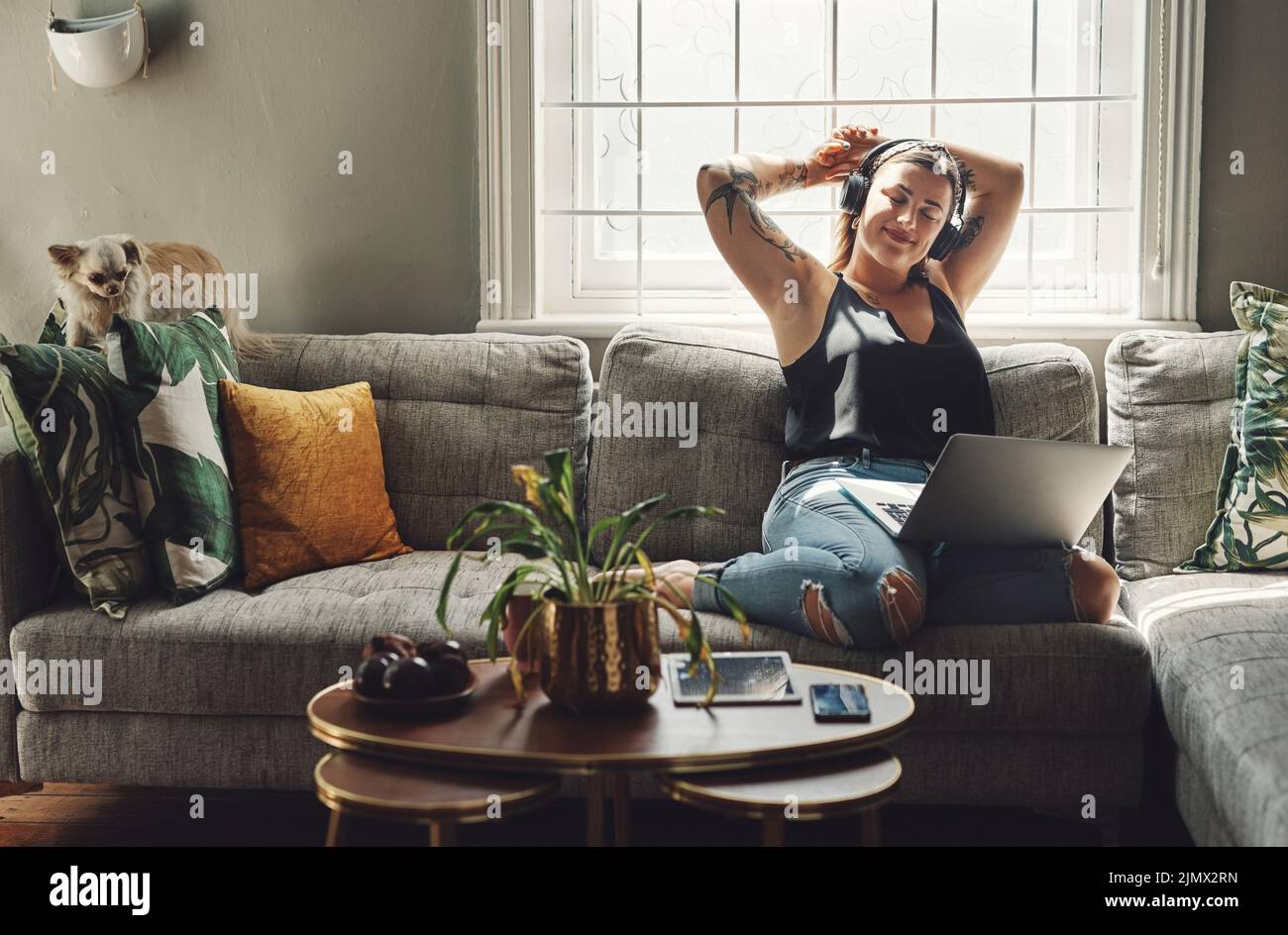 Meine eigene Firma ist meine Lieblingsfirma. Eine junge Frau, die zu Hause einen Laptop und Kopfhörer auf dem Sofa benutzt. Stockfoto
