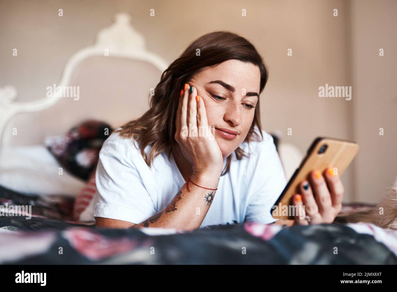 Ich brauche nur etwas, um der Langeweile zu entfliehen. Eine schöne junge Frau benutzt ihr Handy, während sie auf ihrem Bett liegt. Stockfoto