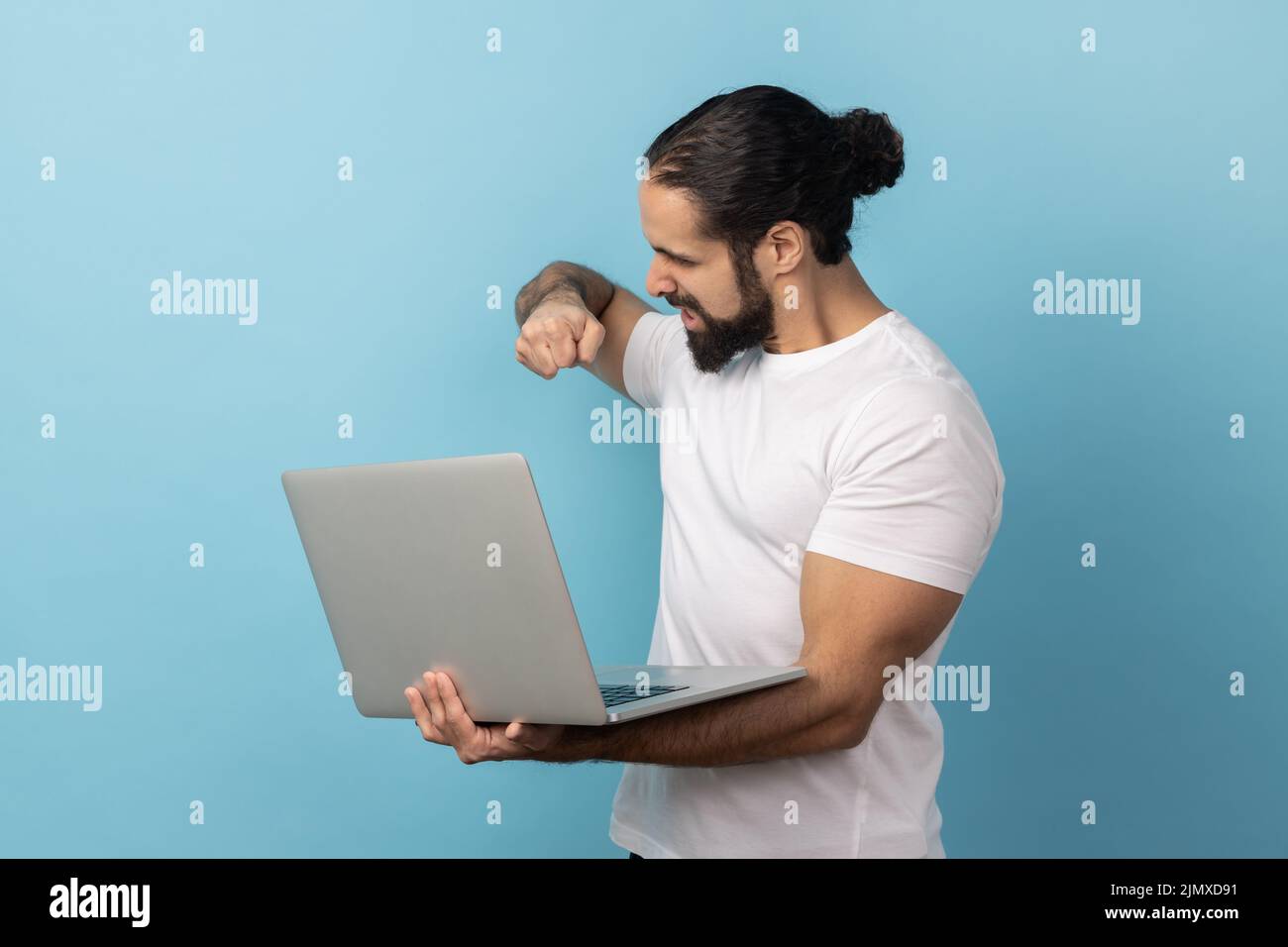 Porträt eines aggressiven Mannes mit Bart in weißem T-Shirt, der am Laptop arbeitet, Fehler hat, die Faust auf den Bildschirm schlagen will. Innenaufnahme des Studios isoliert auf blauem Hintergrund. Stockfoto