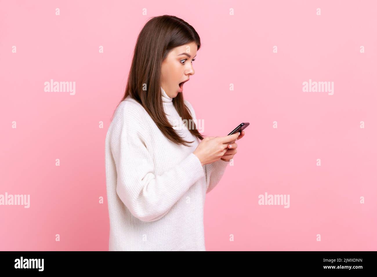 Seitenansicht Porträt der schockierten Bloggerin im Stehen, verwenden Sie Smartphone mit staunendem Gesicht, geöffneten Mund, tragen weißen lässigen Stil Pullover. Innenaufnahme des Studios isoliert auf rosa Hintergrund. Stockfoto