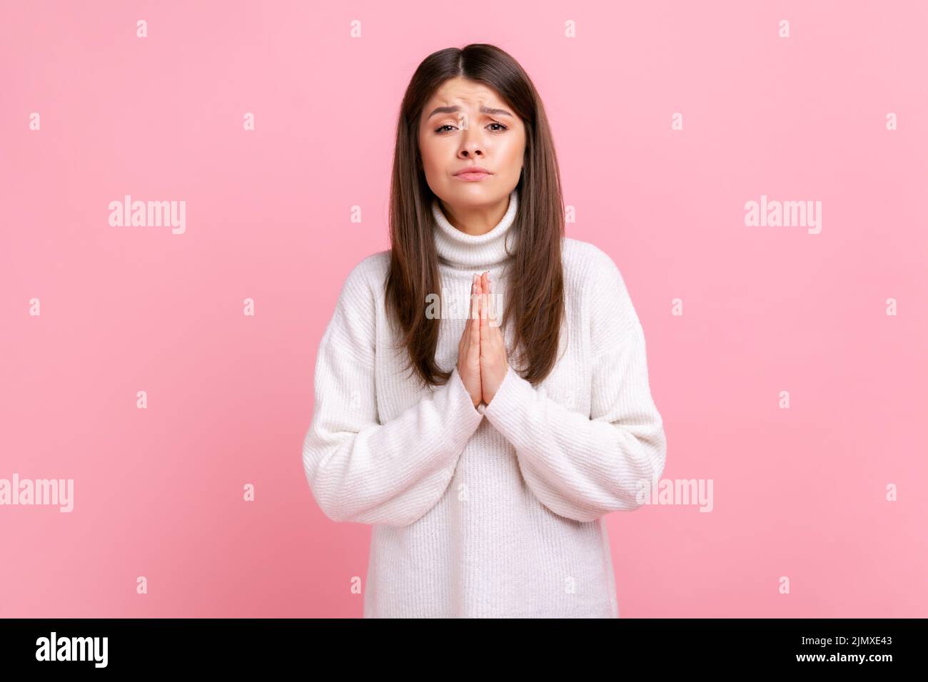 Das Weibchen hält die Hände im Gebet und sieht mit flehenden Bitten, bettelender Hilfe, Vergebung aus, trägt einen weißen Pullover im lässigen Stil. Innenaufnahme des Studios isoliert auf rosa Hintergrund. Stockfoto