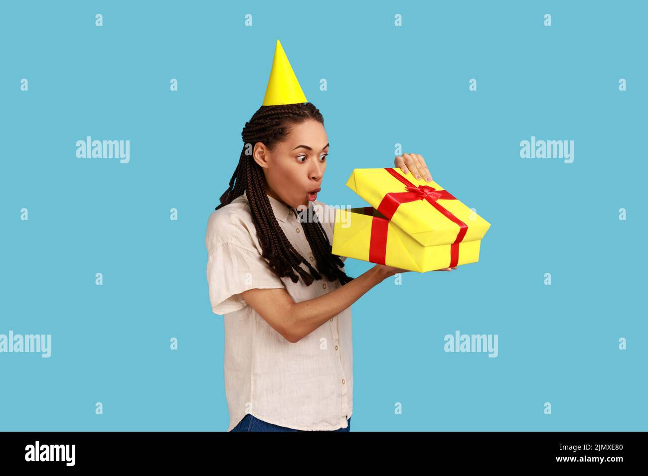 Porträt einer überraschten Frau in gelbem Partykegel, die in die Geschenkbox schaut, das Geschenk öffnet und mit schockierter Ausstrahlung in das Innere blickt, das weißes Hemd trägt. Innenaufnahme des Studios isoliert auf blauem Hintergrund. Stockfoto