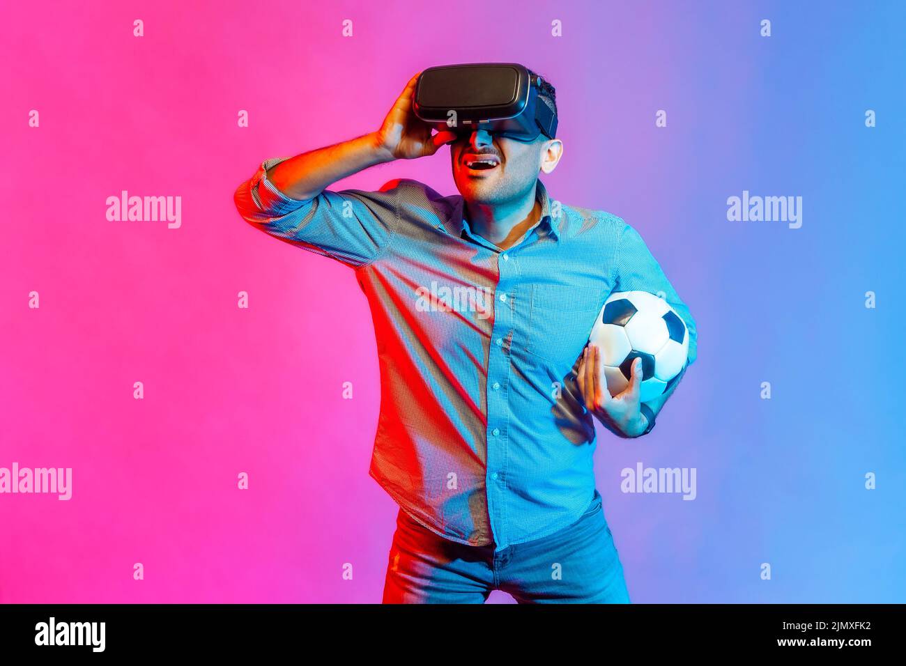 Porträt eines aufgeregten, konzentrierten Mannes im Hemd, der Headset trägt und den Fußballball hält und 3D Fußballmannschaften beobachtet. Innenaufnahmen im Studio, isoliert auf farbigem Neonlicht-Hintergrund. Stockfoto
