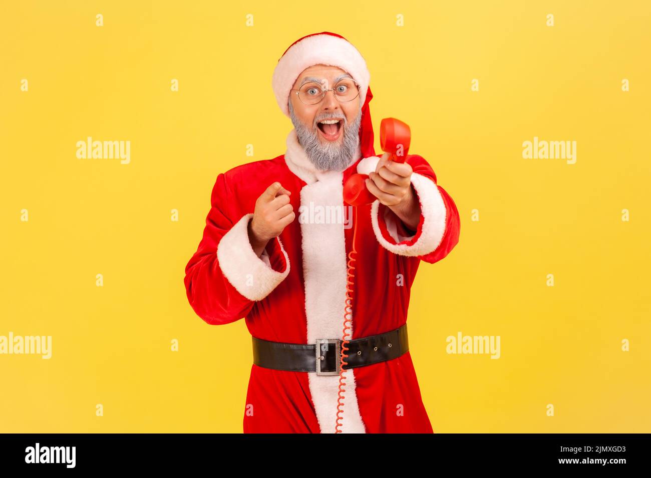 Älterer Mann mit grauem Bart trägt weihnachtsmann Kostüm hält Festnetz-Telefon Hörer, ruft Sie mit Fests zu gratulieren, zeigt auf Kamera. Innenaufnahme des Studios isoliert auf gelbem Hintergrund Stockfoto