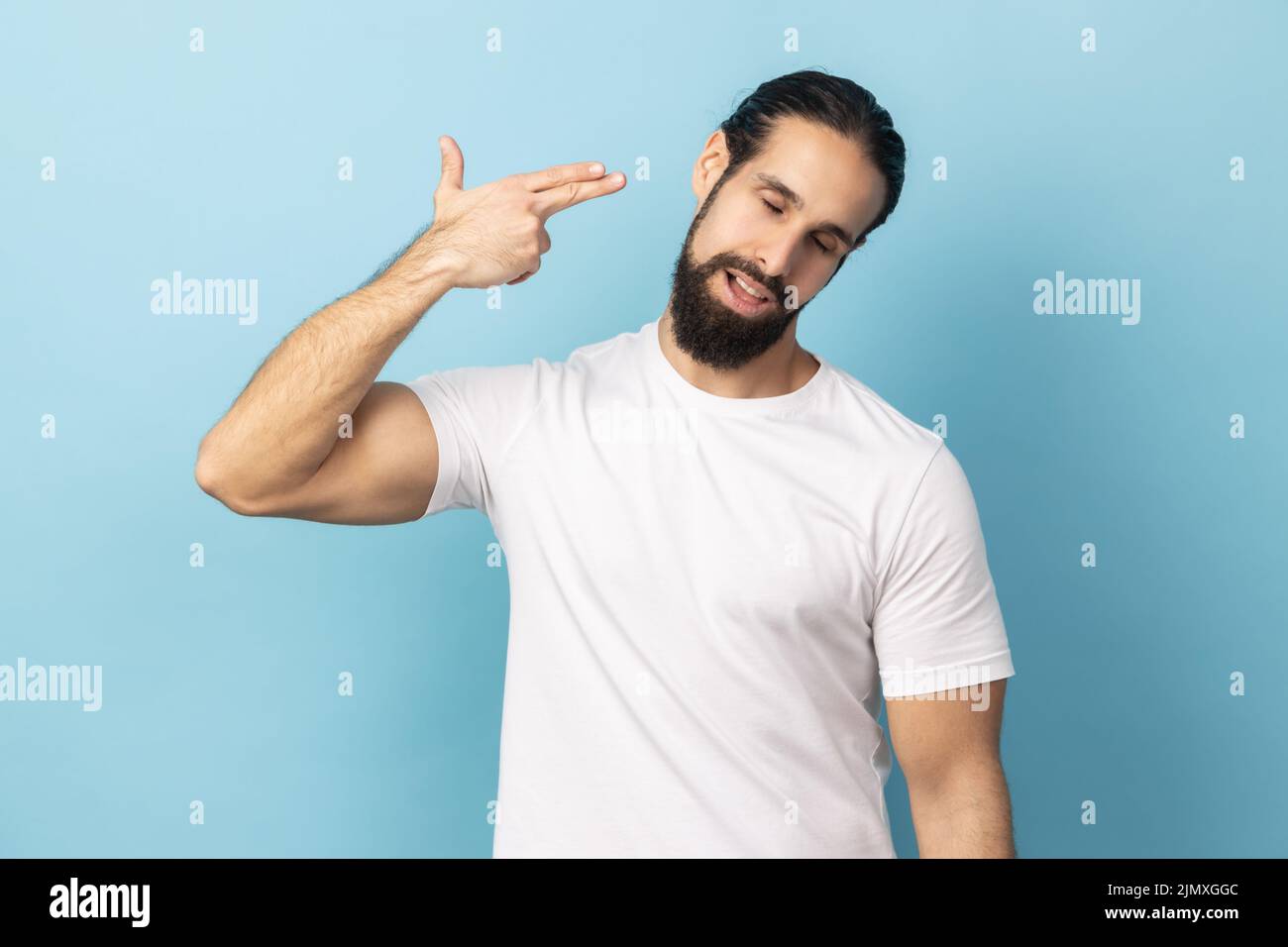 Kopfschuss, tötet mich bitte. Deprimierter Mann mit Bart und weißem T-Shirt, der mit Fingergewehrgesten Selbstmord begeht, verzweifelter Gesichtsausdruck. Innenaufnahme des Studios isoliert auf blauem Hintergrund. Stockfoto