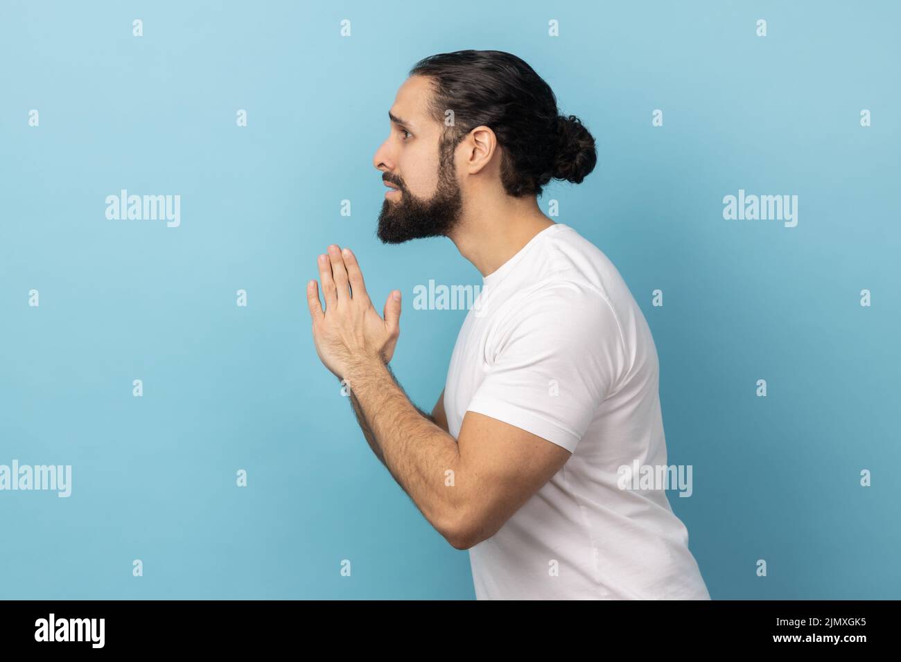 Bitte, ich bitte um Verzeihung. Seitenansicht eines verärgerten Mannes mit Bart und weißem T-Shirt, der mit verzweifelter Grimasse um Hilfe betet. Innenaufnahme des Studios isoliert auf blauem Hintergrund. Stockfoto