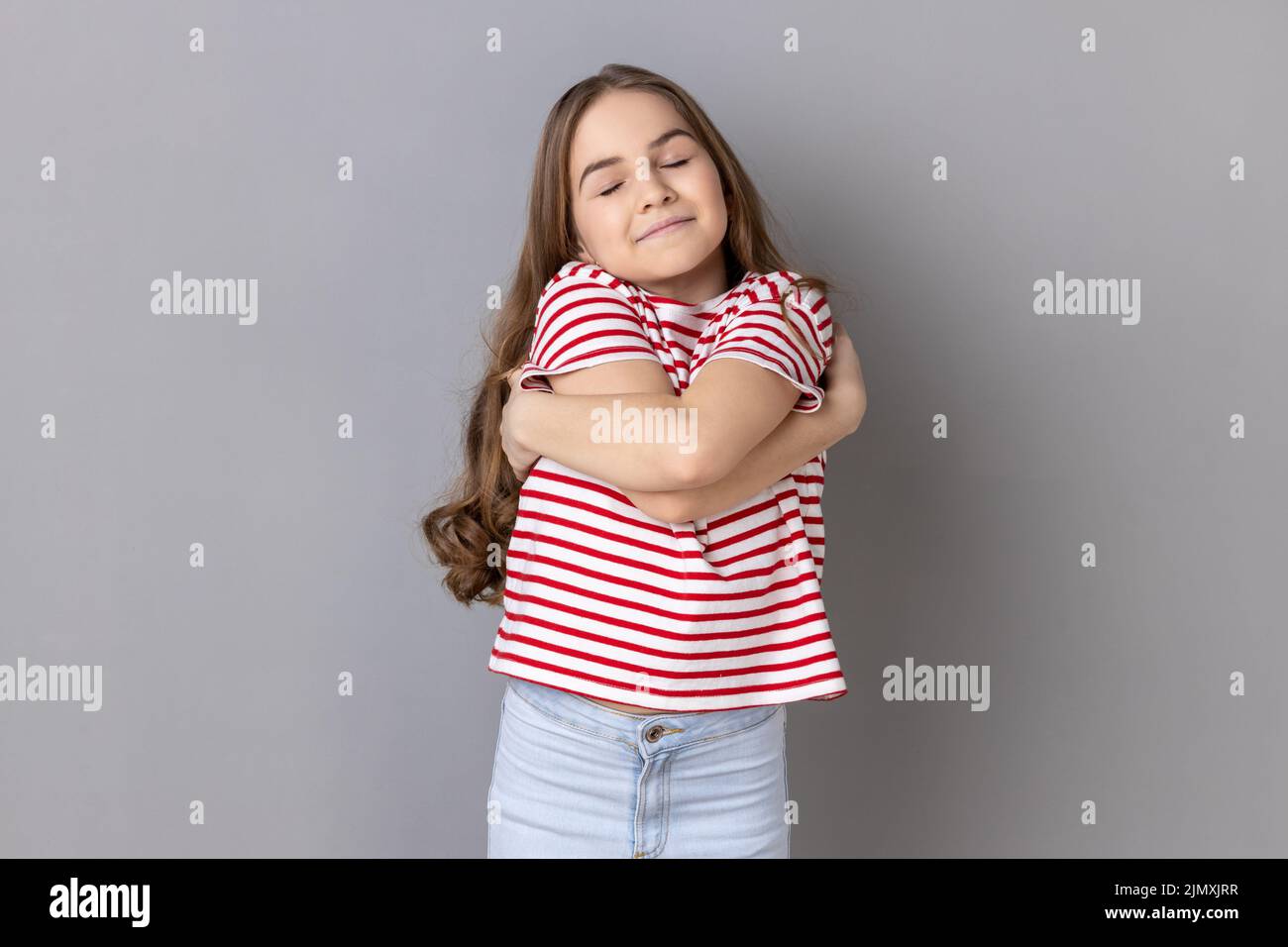 Ich liebe mich. Porträt eines selbstzufriedenen egoistischen kleinen Mädchens mit gestreiftem T-Shirt, das sich umarmt und mit Freude lächelt und sich selbststolz fühlt. Innenaufnahme des Studios isoliert auf grauem Hintergrund. Stockfoto