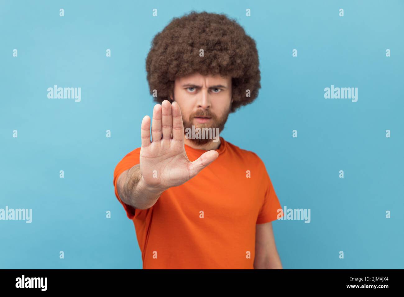 Porträt eines Mannes mit Afro-Frisur in orangefarbenem T-Shirt mit Stop-Geste mit Handfläche, Konfliktverbotswarnung vor Gefahr, Mobbing stoppen. Innenaufnahme des Studios isoliert auf blauem Hintergrund Stockfoto