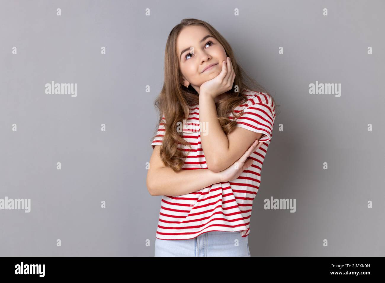 Porträt eines glücklichen positiven kleinen Mädchens mit gestreiftem T-Shirt, das mit der Hand unter dem Kinn steht und träumt und lächelnd wegschaut. Innenaufnahme des Studios isoliert auf grauem Hintergrund. Stockfoto