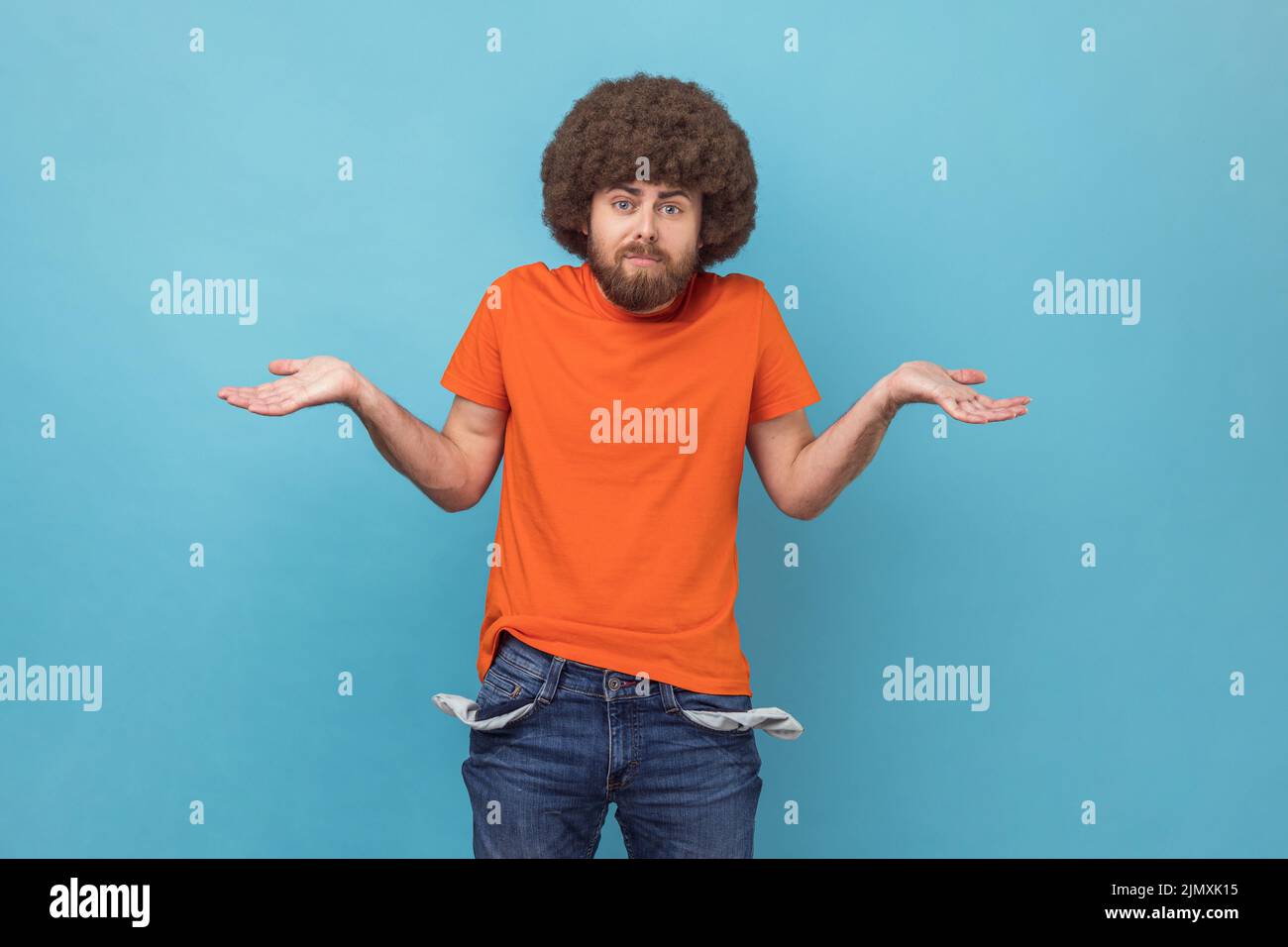 Porträt eines Mannes mit Afro-Frisur in orangefarbenem T-Shirt mit leeren Taschen und frustrierten Blick über Kredite und Schulden, hat kein Geld, arbeitslos. Innenaufnahme des Studios isoliert auf blauem Hintergrund. Stockfoto
