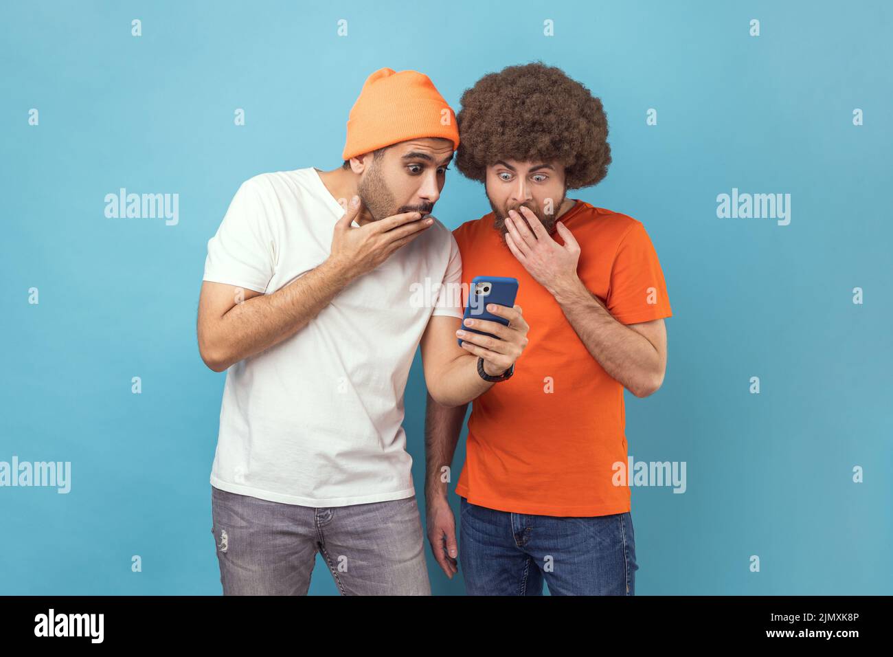 Porträt von zwei attraktiven schockierten jungen erwachsenen Hipster-Männern, die mit dem Smartphone stehen, in sozialen Netzwerken aktuelle Nachrichten lesen und dabei offenen Mund decken. Innenaufnahme des Studios isoliert auf blauem Hintergrund. Stockfoto