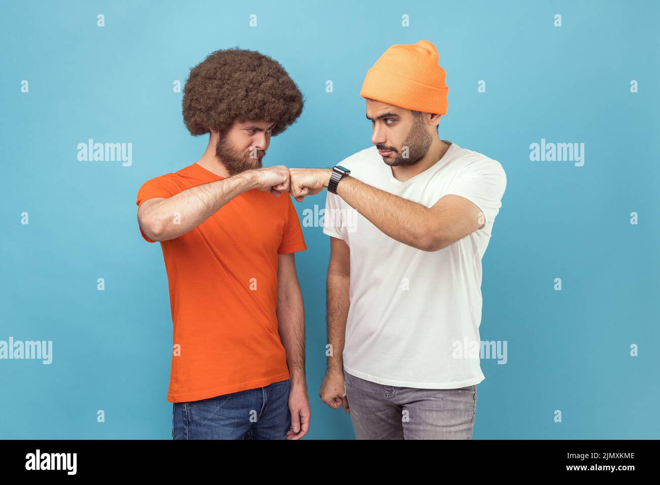 Portrait von zwei ernsthaften jungen erwachsenen Hipster-Männern, die sich mit aggressiver Optik und mit Beule-Geste im Kampf angucken und stehen. Innenaufnahme des Studios isoliert auf blauem Hintergrund. Stockfoto