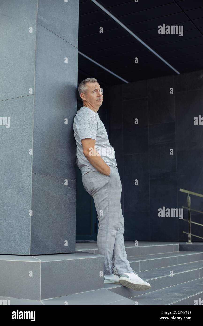 Porträt eines müden grauen Geschäftsmannes, der sich auf die graue Konstruktion eines modernen Bürogebäudes in der Nähe des Eingangs stützt. Urbaner Lifestyle, stylisch Stockfoto
