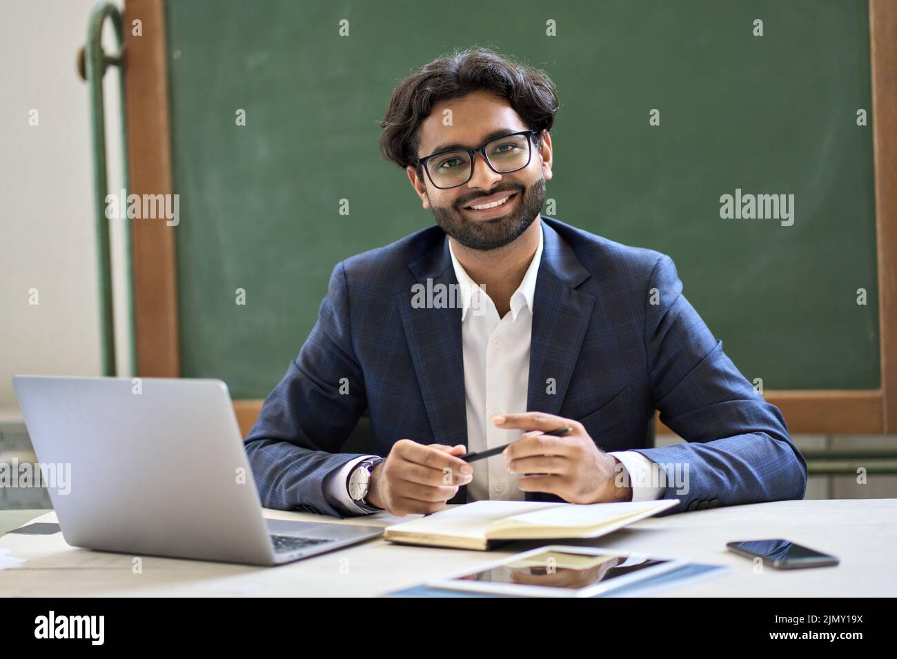 Glücklicher junger indischer Geschäftsmann Lehrer sitzt bei der Arbeit im Klassenzimmer. Hochformat Stockfoto