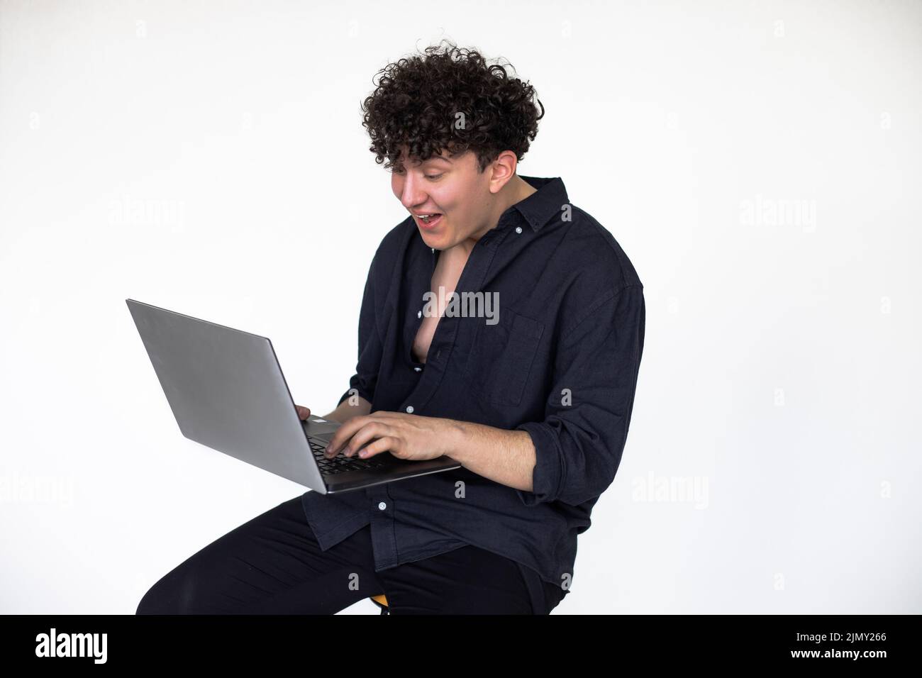 Junger lockiger Kerl in total schwarzem Look, der auf einem Stuhl sitzt und Computerspiele auf einem modernen Laptop spielt, grauer Studiohintergrund. Spieleinnahme Stockfoto