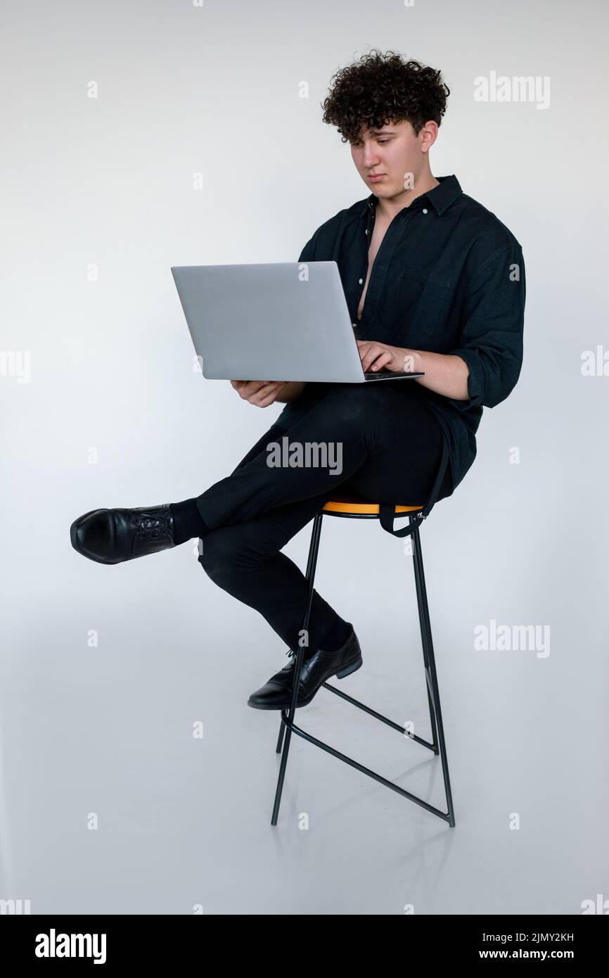 In voller Länge Foto von verärgert jungen lockigen Kerl in total schwarz aussehen sitzen mit gekreuzten Beinen auf dem Stuhl und spielen Computerspiele auf einem modernen Laptop, grau Studio Stockfoto