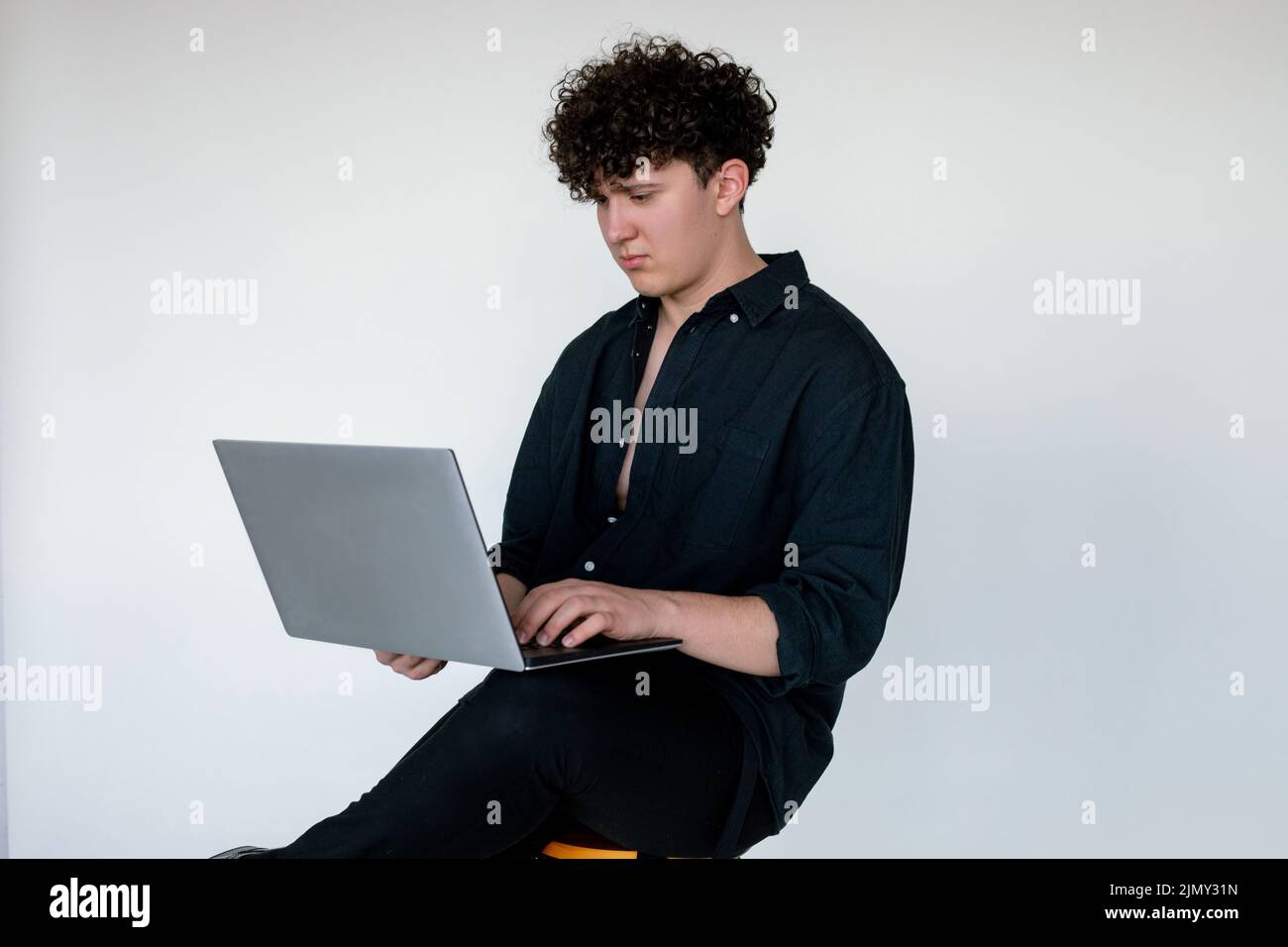 Hübscher junger lockiger Kerl in total schwarzem Look, der auf einem Stuhl sitzt und Computerspiele auf einem modernen Laptop spielt, grauer Studiohintergrund. Absorption in Stockfoto