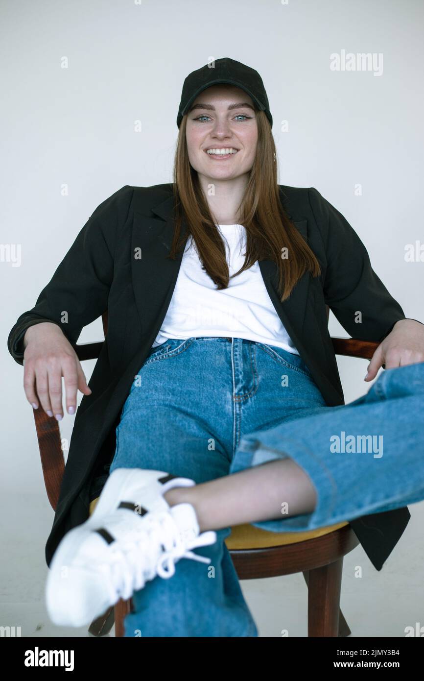 Porträt eines lächelnden Mädchens auf einem Stuhl sitzend, grauer Studiohintergrund, Kopierraum. Lässige schwarze Blazer und Mütze für Frauen, Jeans und Gummischuhe. Komfortabel Stockfoto