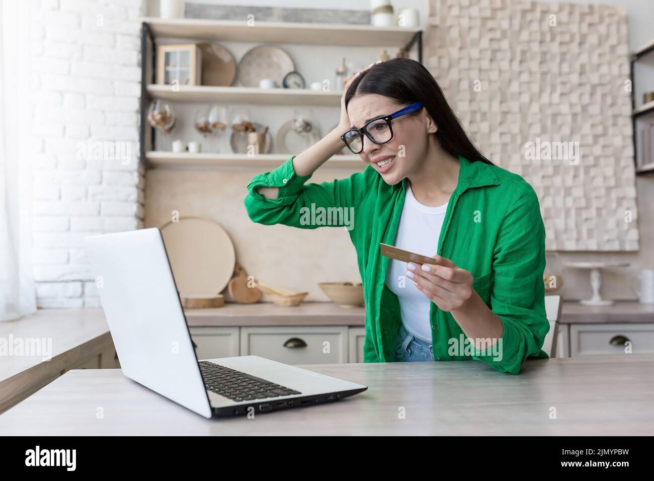 Währungskrise. Kreditschuld. Verärgert junge Frau in Brille und grünem Hemd mit Kreditkarte, sitzt am Laptop zu Hause, hält den Kopf, schockiert, verwirrt. Stockfoto