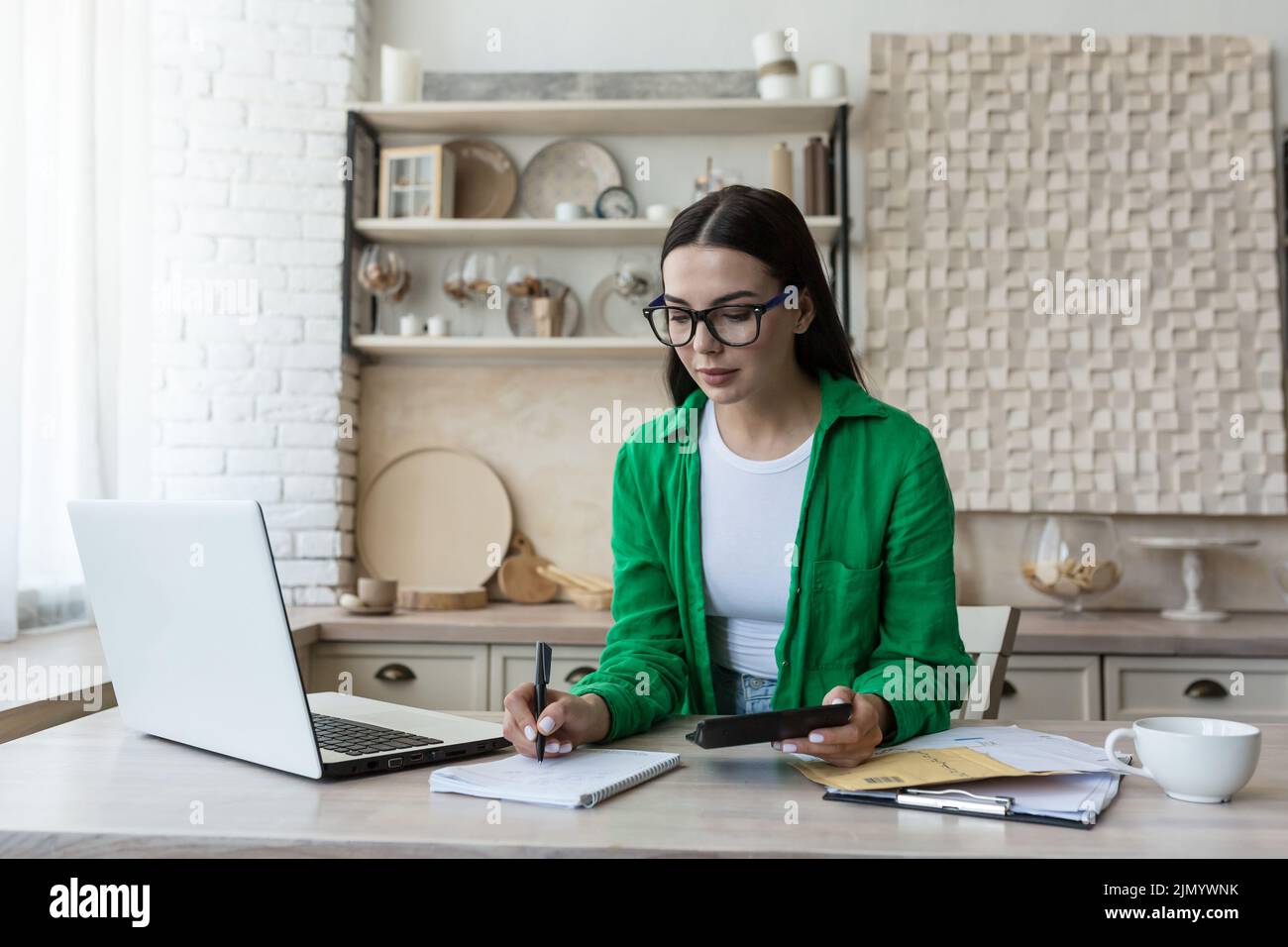 Hausbuchhaltung.Eine junge Frau zählt ihr Einkommen und expenses.checks das Haushaltsbudget. In einer Brille und einem grünen Hemd, sitzt zu Hause mit einem Laptop, einem Rechner und schreibt in einem Notebook, berechnet. Stockfoto
