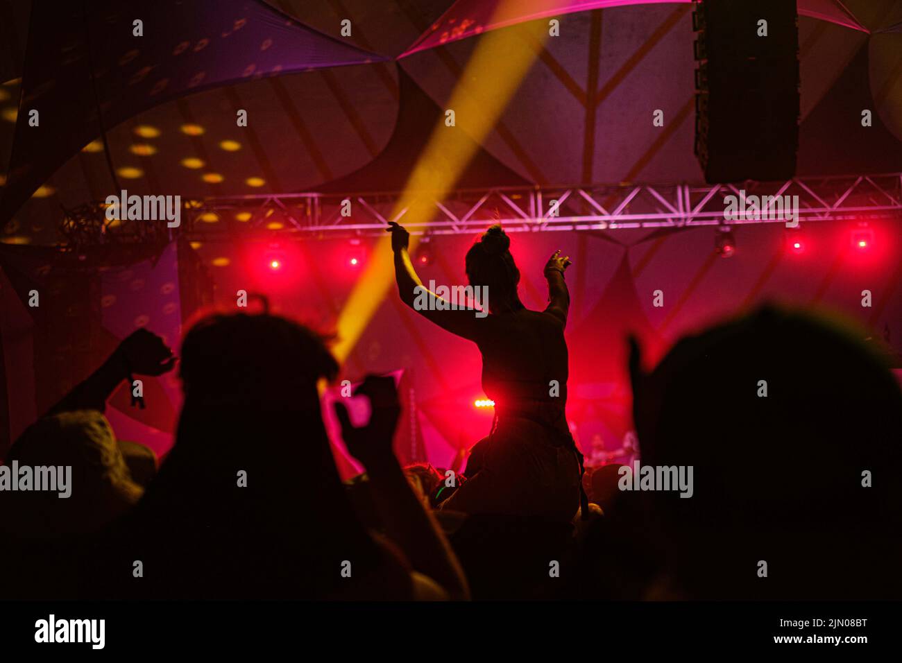 Eine Silhouette einer jungen Frau, die beim Latitude Music Festival in Suffolk unter rosa Lichtern auf den Schultern ihres Freundes sitzt Stockfoto