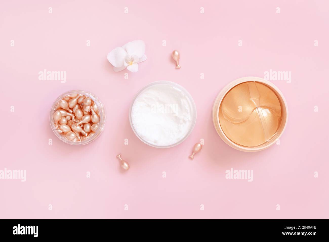 Gesicht Serumkapseln, Creme und Augenflecken auf hellrosa in der Nähe von Orchideenblüte Draufsicht. Hautpflegeprodukte. Schönheitsbehandlungen im Gesicht. Pastell minimal com Stockfoto