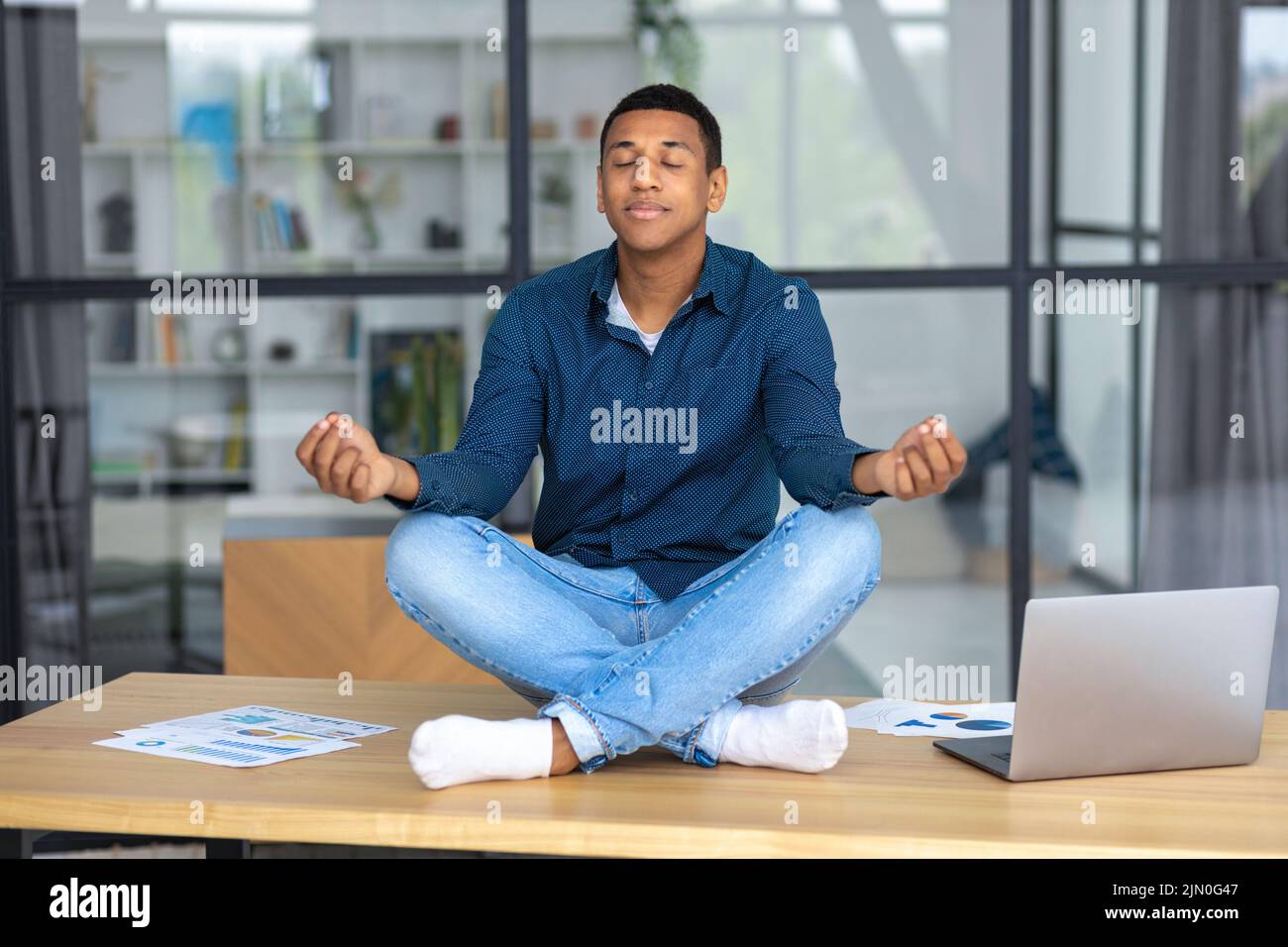 Ruhiger junger Mann, der am Arbeitsplatz im Büro sitzt und meditiert, eine Pause zu machen und stressigen Job zu vermeiden Stockfoto