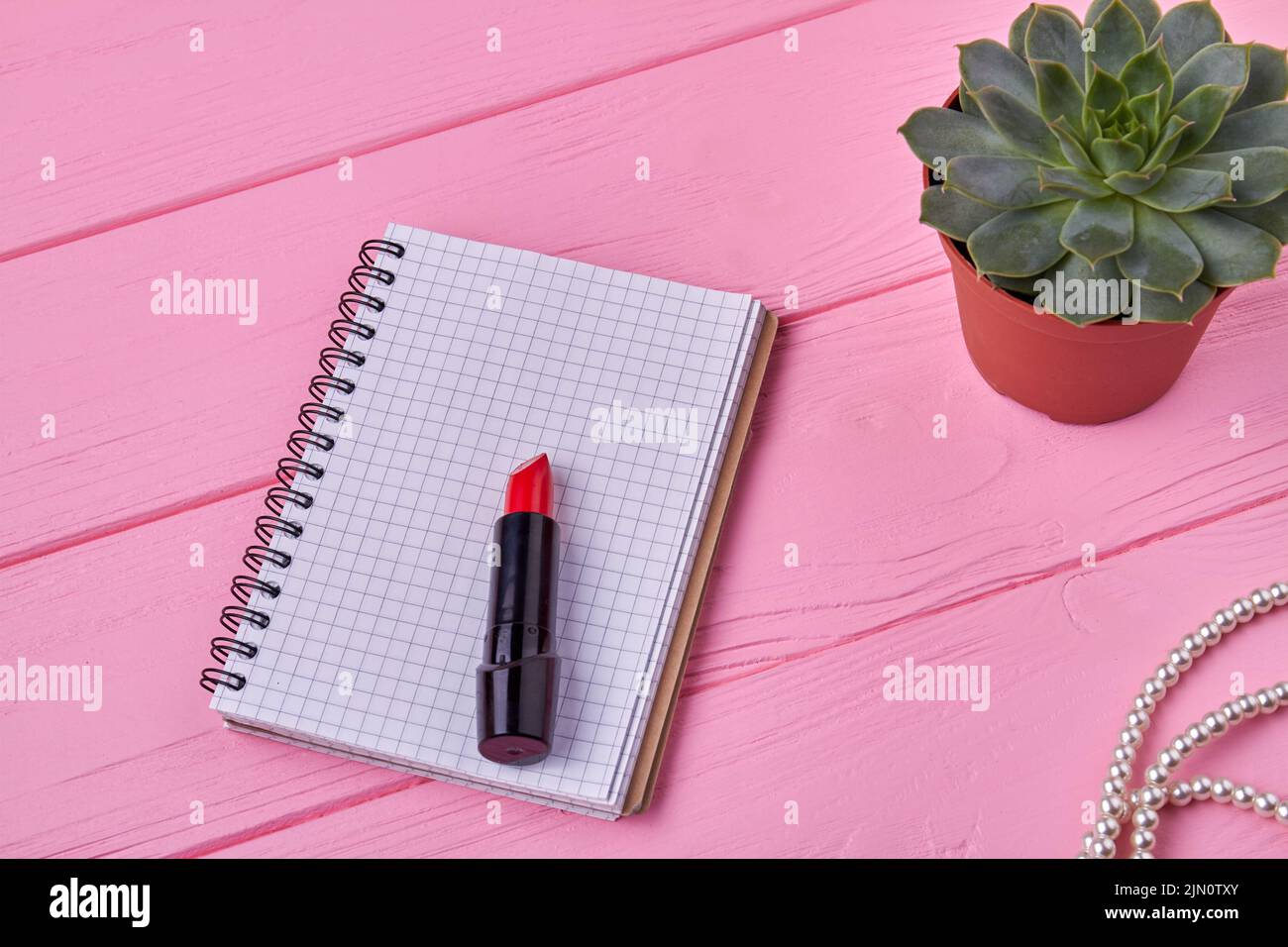 Notizblock von oben mit Lippenstift auf rosafarbenem Holzschreibtisch. Notizbuch mit Kopierplatz. Stockfoto