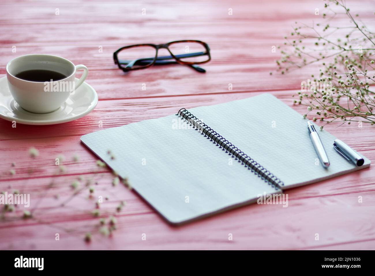 Notizblock mit Kaffeetasse und Gläsern auf rosafarbenem Holzschreibtisch. Bouquet von getrockneten Frühlingsblumen. Stockfoto
