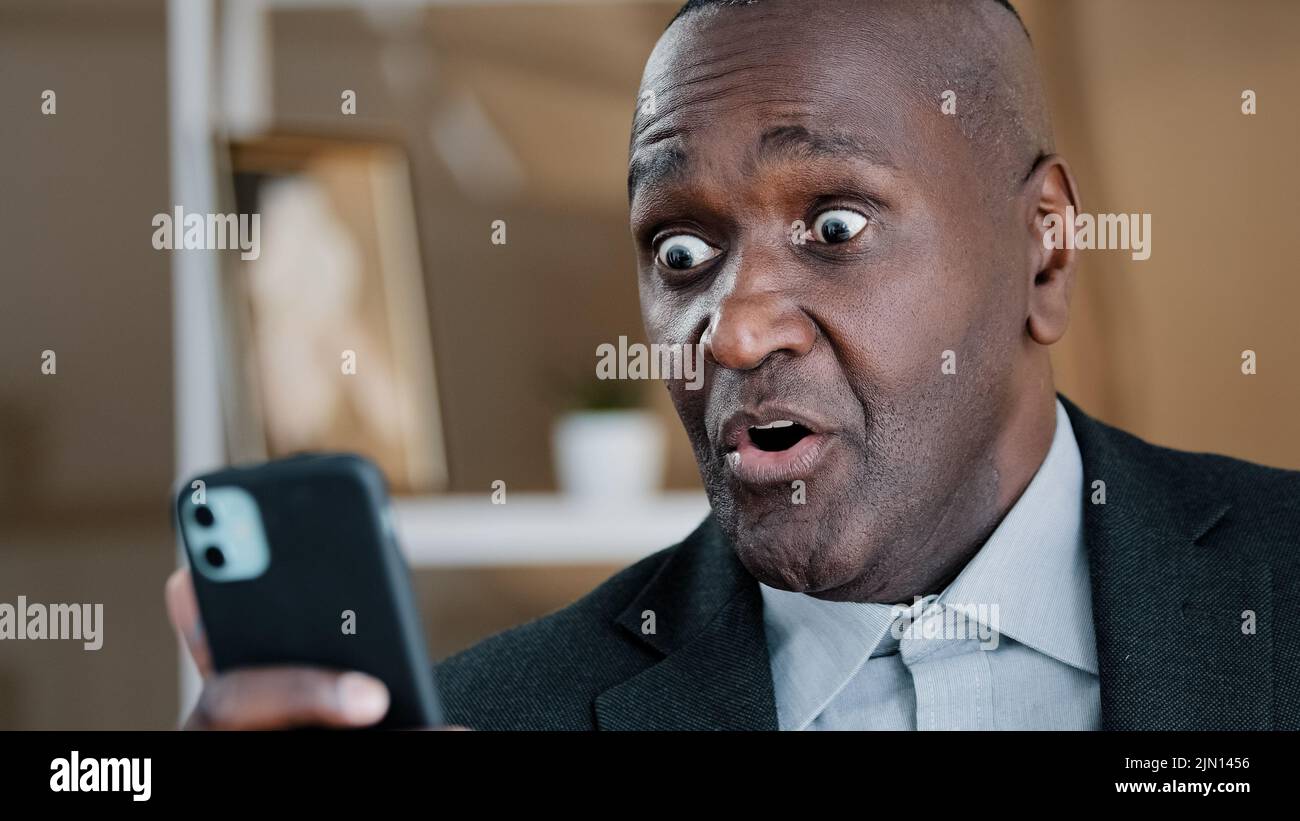 Der afroamerikanische alte 50s überraschte den schockierten Mann, der auf den Bildschirm des Mobiltelefons schaute und unglaubliche Nachrichten las, die mit dem Kopf winkten, ohne Meinungsverschiedenheiten zu negieren Stockfoto
