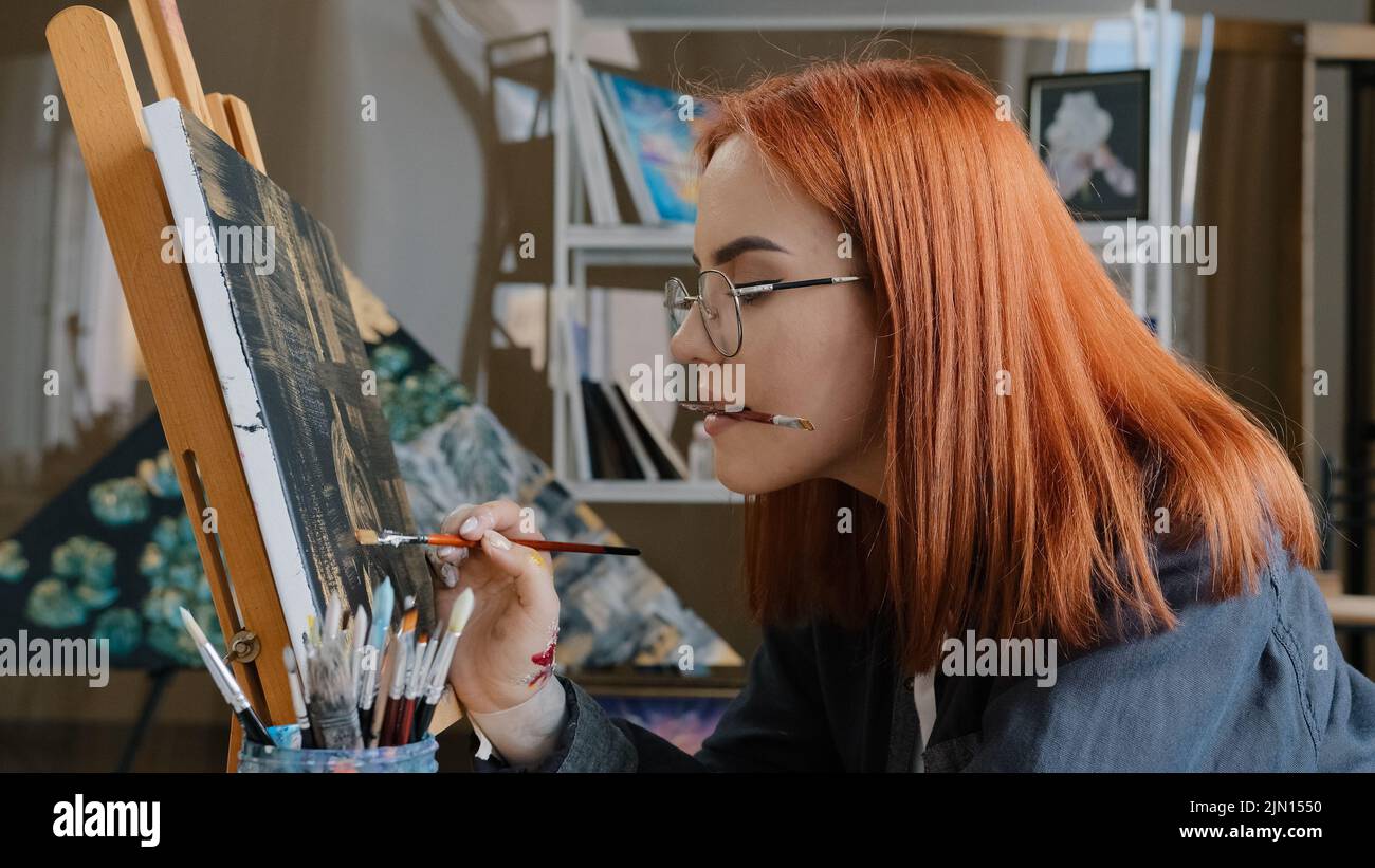 Inspiriert lustig fokussierte Künstlerin Frau mit roten Haaren Mädchen Maler trägt Gläser hält Malerei Pinsel in Mund Farben mit Ölfarben auf Leinwand auf Staffelei Stockfoto