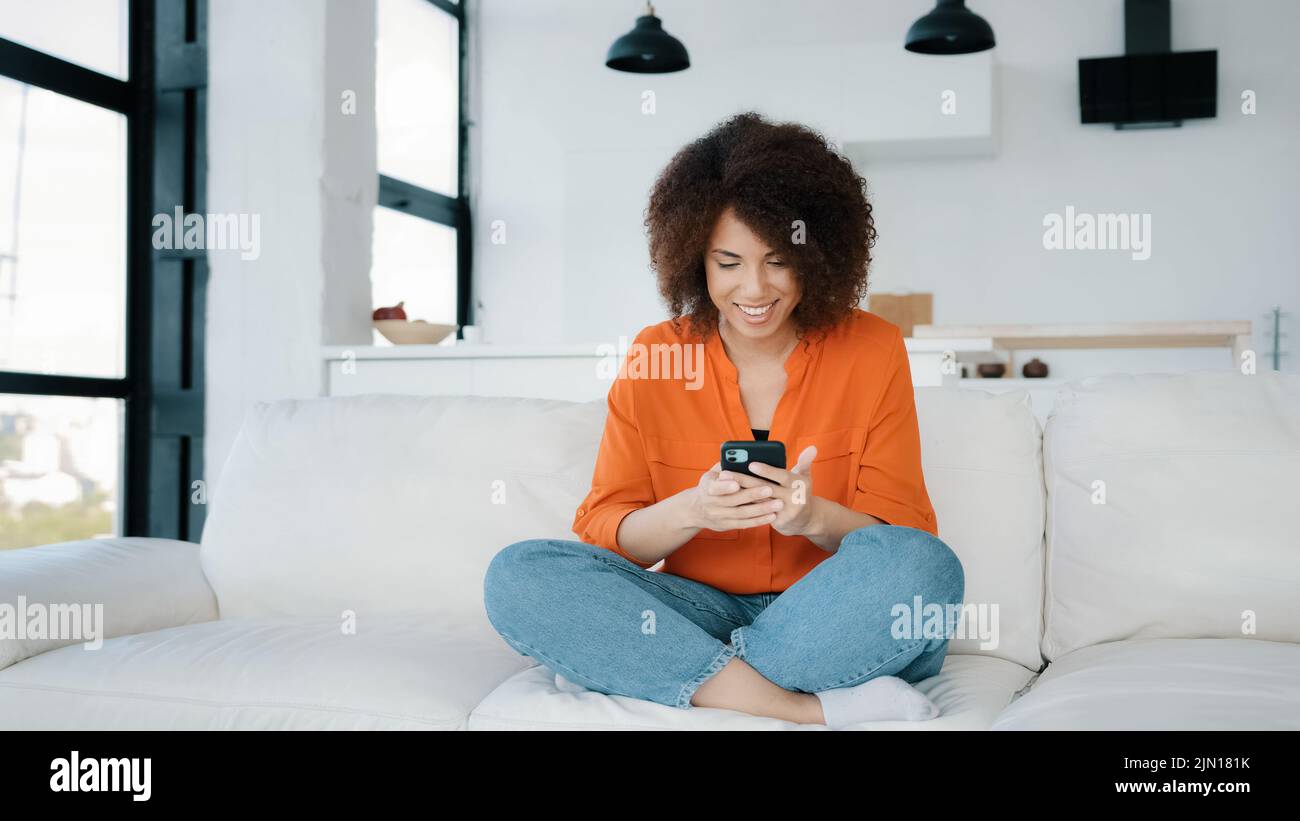 Afroamerikanische Frau sitzt zu Hause Couch suchen Smartphone mit Handy spielen mobile Spiel Sieg Reaktion erhalten gute Angebot Gelegenheit Nachrichten Stockfoto