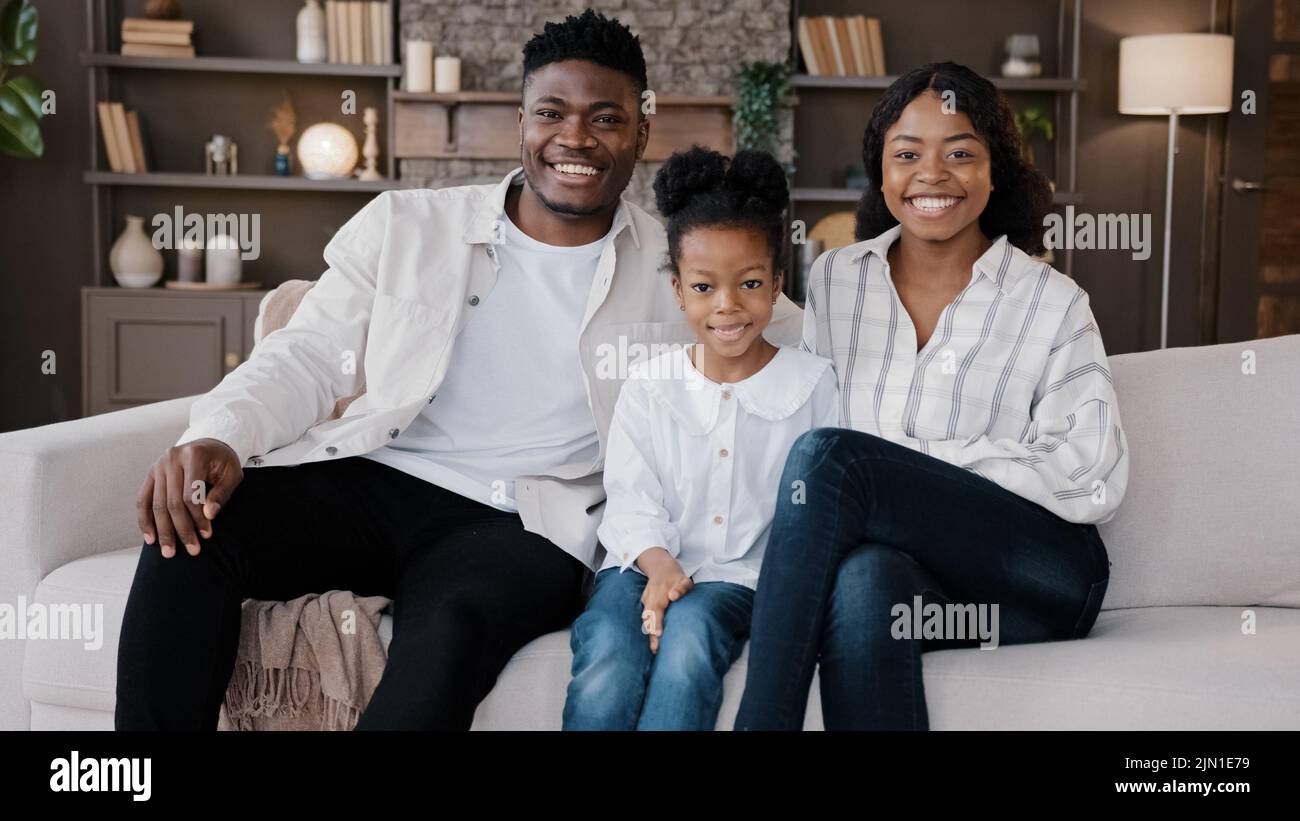 Afrikanische Familie setzt sich auf eine komfortable Couch im Wohnzimmer. Biracial Eltern und Kind lächeln Blick auf die Kamera. Fröhliche Tochter mit Mama und Papa Stockfoto