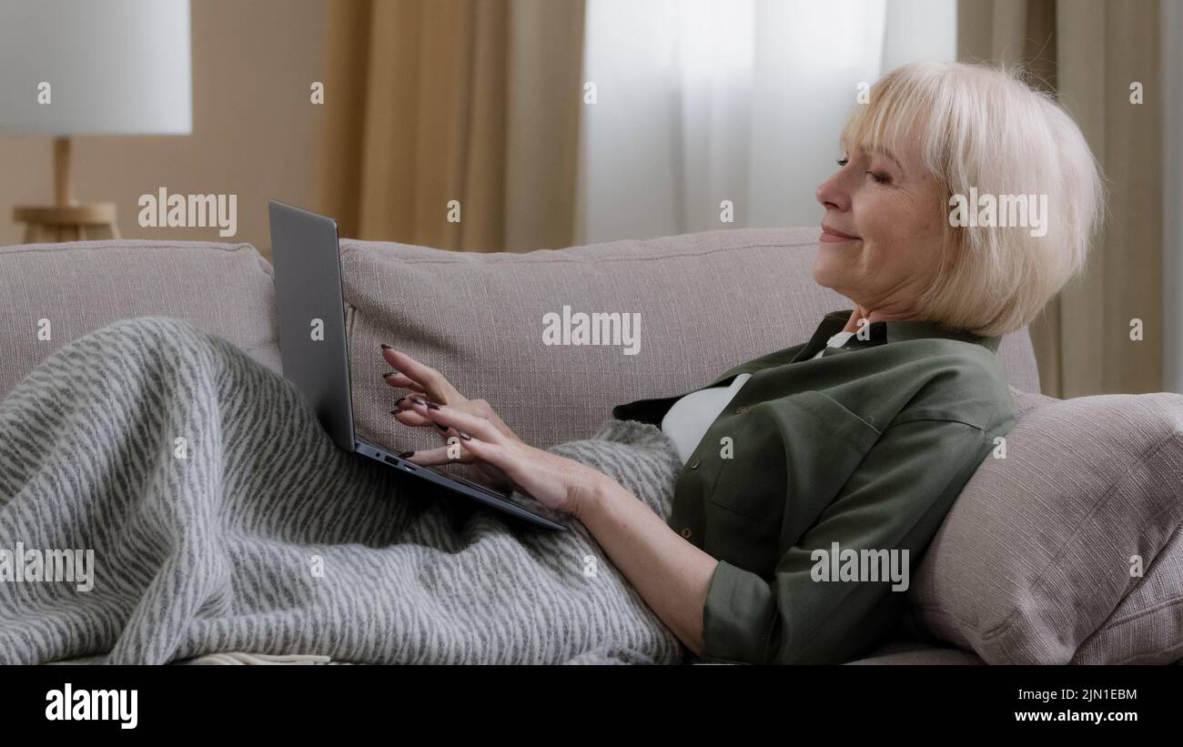 Alte ältere kaukasische grauhaarige Frau sitzt auf der Couch, die mit einer Decke bedeckt ist, die Laptop schreibt E-Mail in der Web-Computer-Anwendung. Ältere reife Frau bei Stockfoto