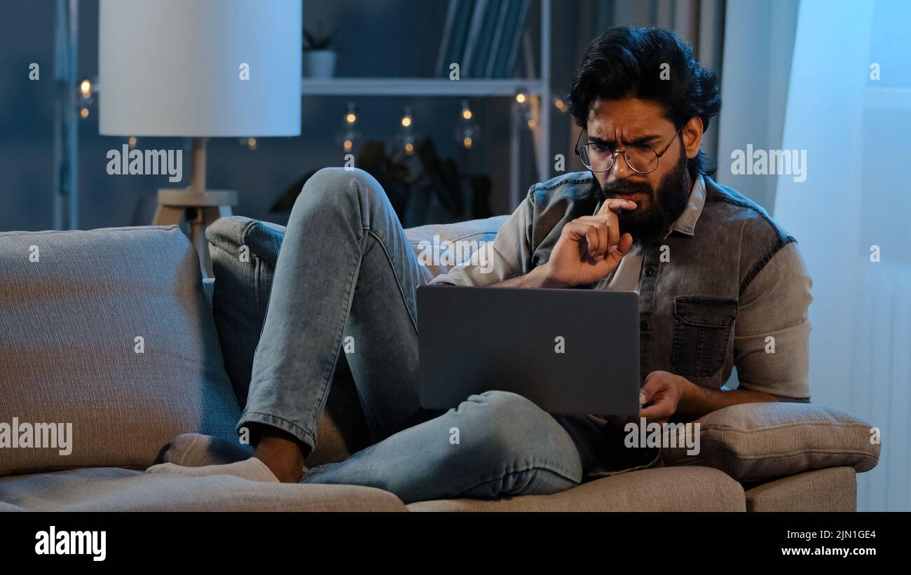 Geschäftsmann arabischer indischer hispanischer Mann bärtiger Benutzer, der eine Brille trägt und nachts auf dem Sofa sitzt, mit Laptop-Computer-Schwierigkeiten bei Online-Problemen Stockfoto