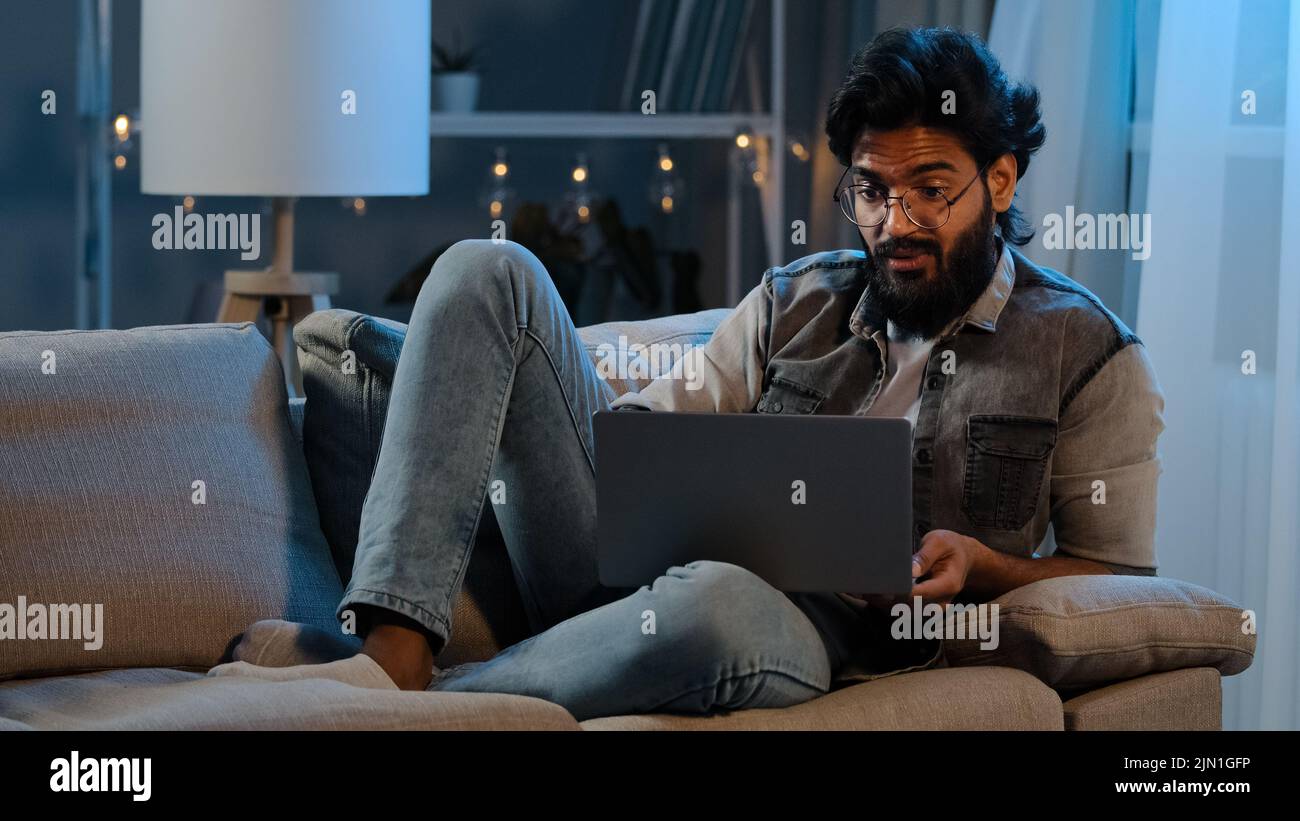 Geschäftsmann Kerl Arab Inder Mann bärtig männlichen Benutzer trägt Brille sitzen auf dem Sofa in der Nacht spät mit Laptop lesen schlechte Nachrichten online Trouble App Stockfoto