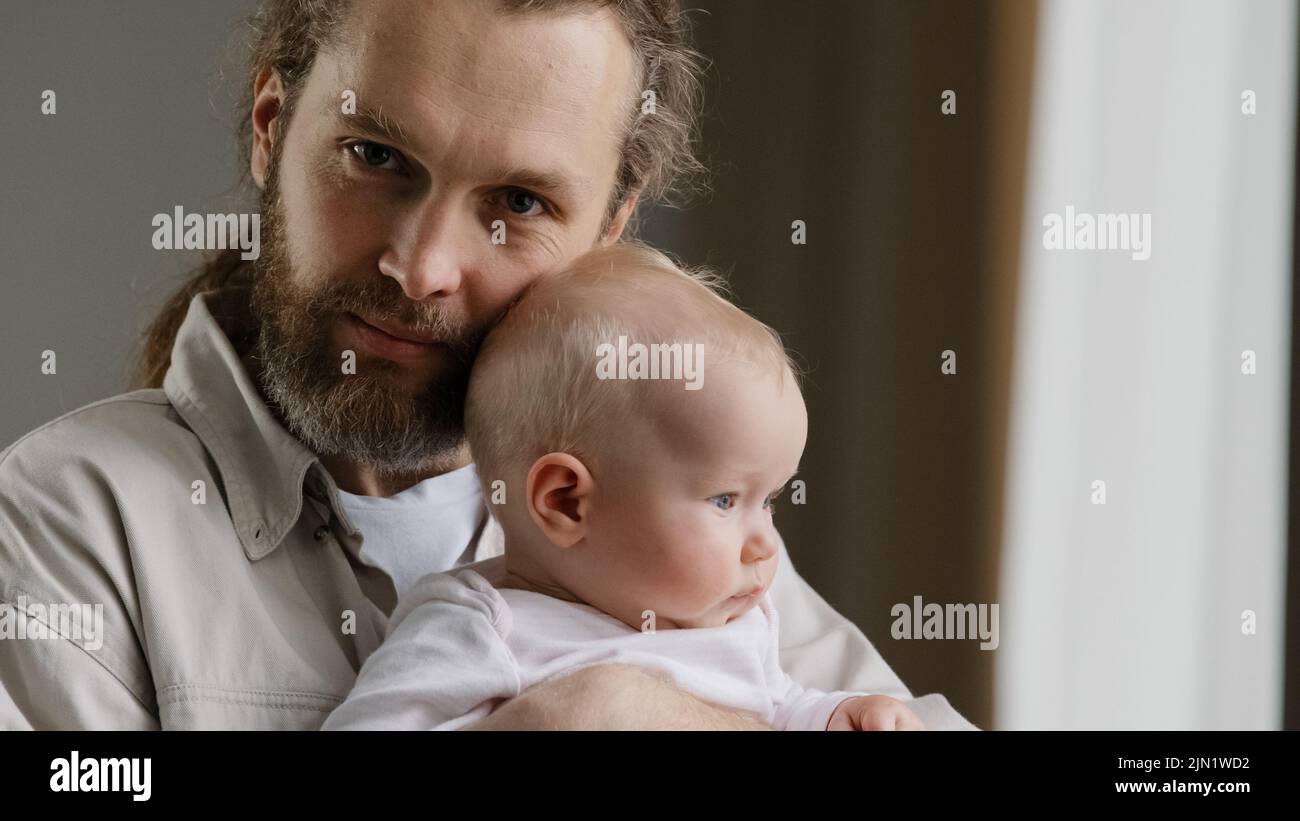 Familienportrait seriöse kaukasische bärtige Vater Vater Mann hält umarmt kleinen Säugling neugeborenes Mädchen Junge Kind Tochter Sohn Kuss auf den Kopf Blick auf die Kamera Stockfoto