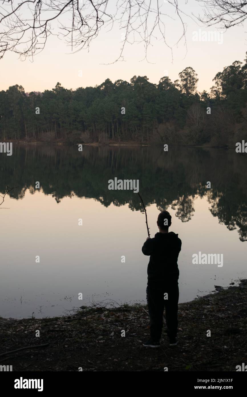 Mann, der in der Abenddämmerung mit einem Baumzweig in einem See im Wald angeln wird Stockfoto