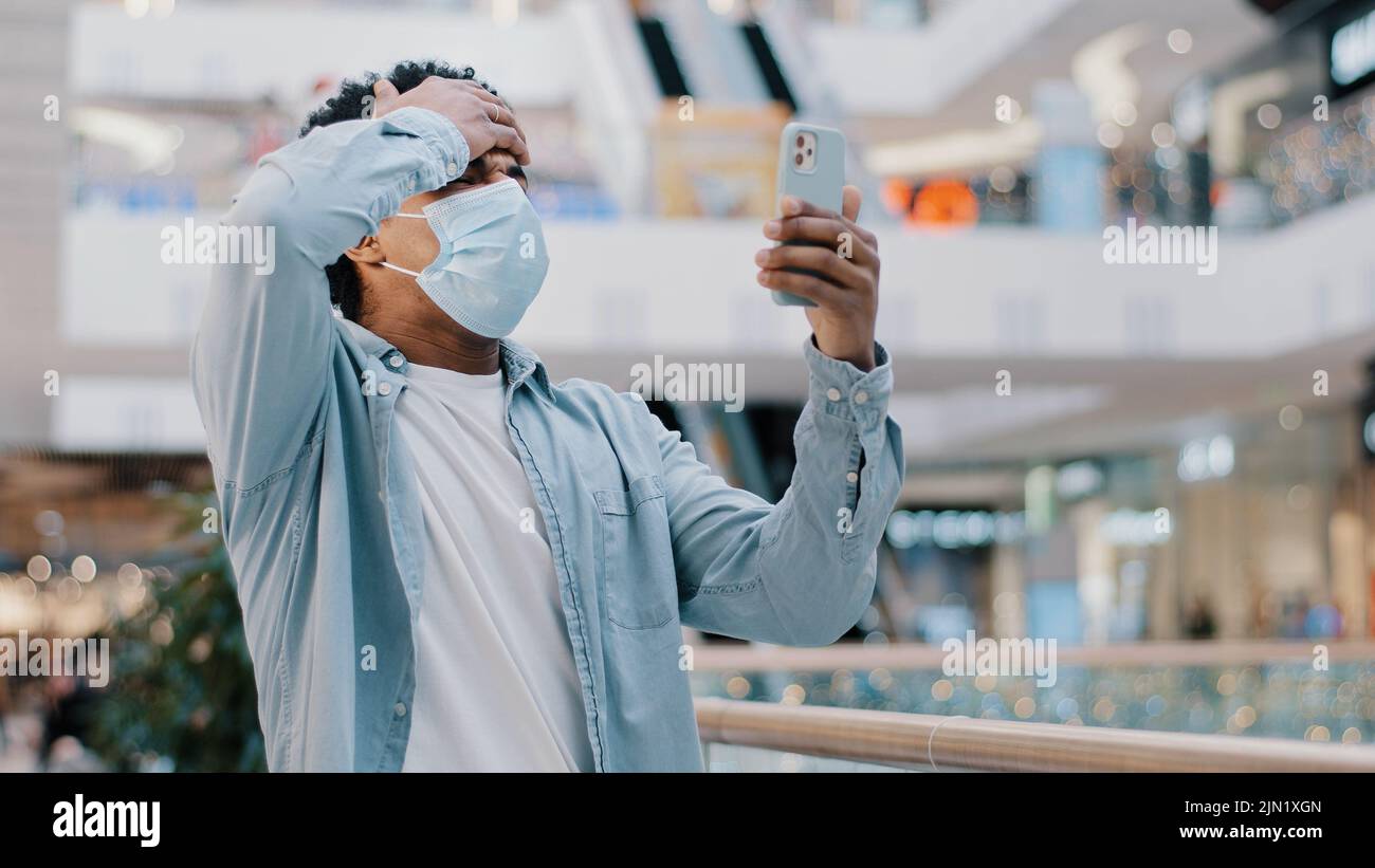 afroamerikanischer Mann mit medizinischer Schutzmaske in Innenräumen Quarantäne-covid Pandemie Blick auf Handy verloren Verlust empfangenden Nachricht Ablehnung Stockfoto