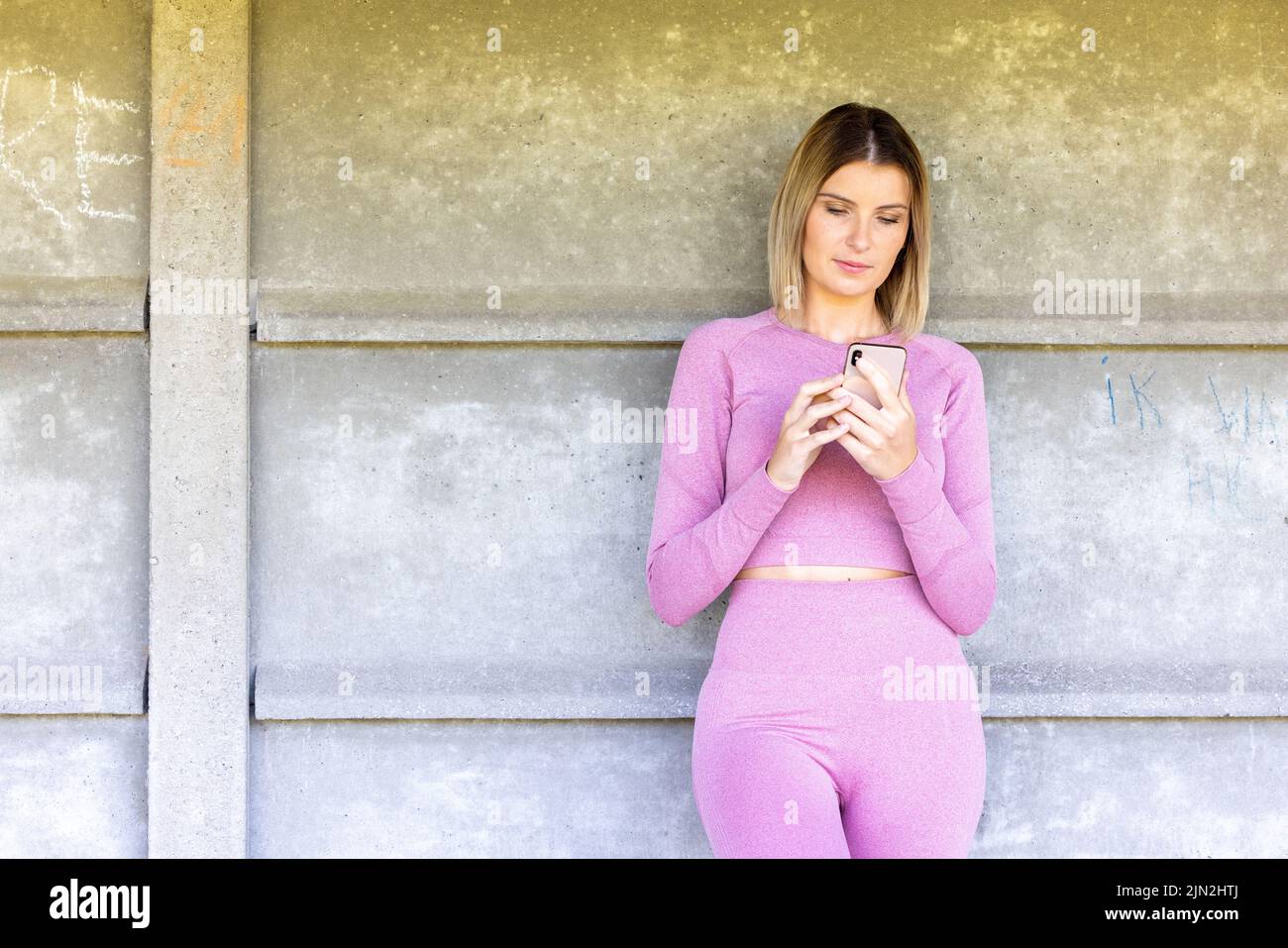 Ganzkörper-Foto einer jungen blonden kaukasischen sportlichen Frau trägt rosa Sportkleidung nutzt Handy im Fitnesscenter. Frau spielen Spiel auf dem Smartphone im Fitnessstudio. Sportliche Mädchen sozialen Netzwerken mit Smartphone im Fitnessstudio nach dem Training. Hochwertige Fotos Stockfoto