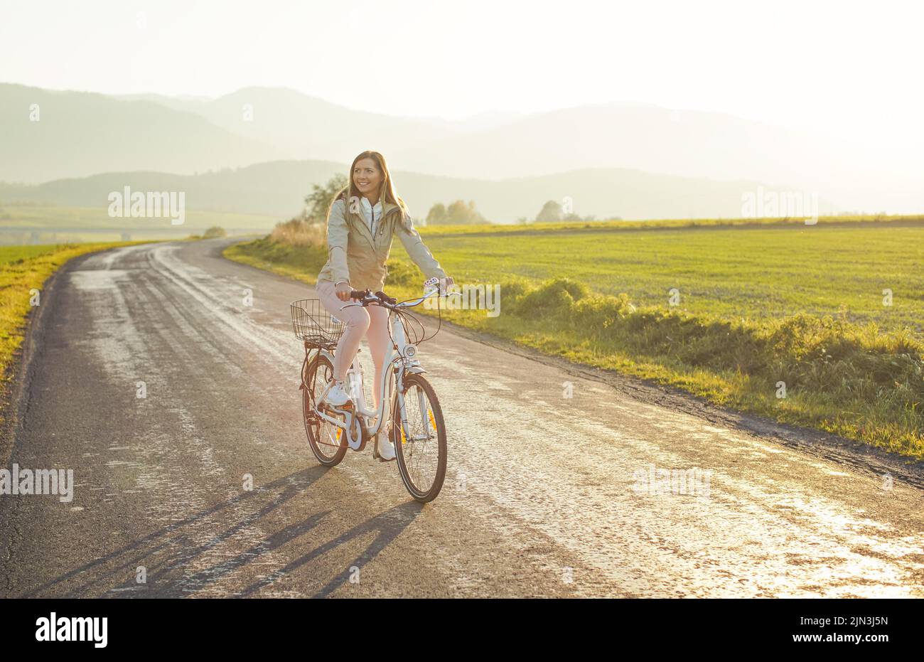 Eine junge Frau in Jacke fährt mit dem Fahrrad über die Landstraße, die Nachmittagssonne scheint im Hintergrund Stockfoto