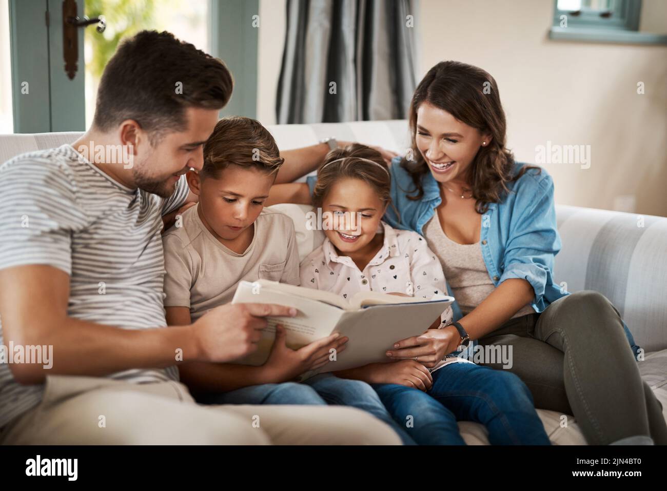 Technologie verbindet uns mit der Familie auf mehr als eine Art und Weise. Eine junge vierköpfige Familie liest zu Hause auf dem Sofa zusammen ein Buch. Stockfoto
