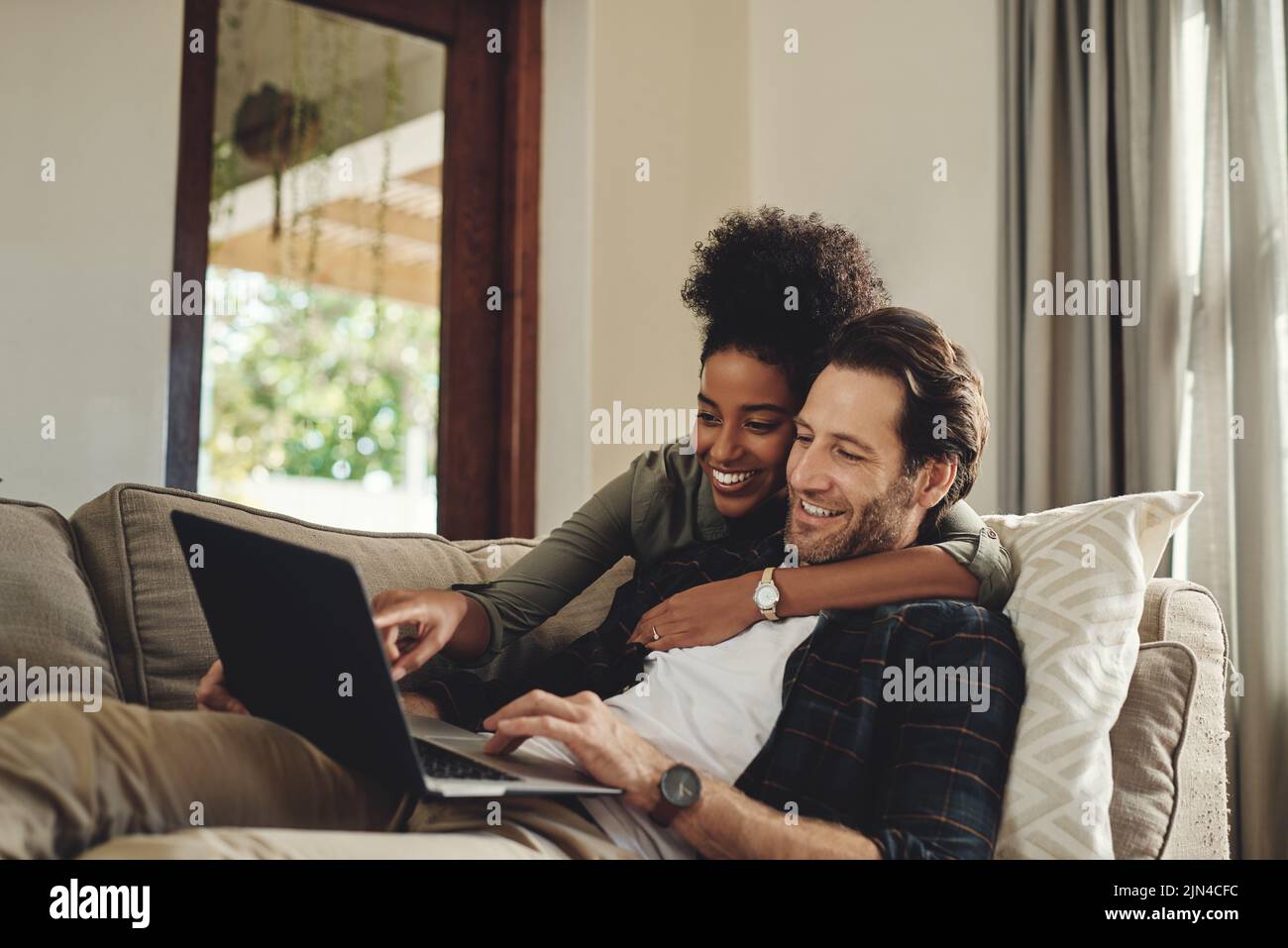 Schauen Sie sich diese Hüne an. Ein glückliches junges Paar, das einen Laptop benutzt, während es sich auf einer Couch im Wohnzimmer zu Hause entspannt. Stockfoto