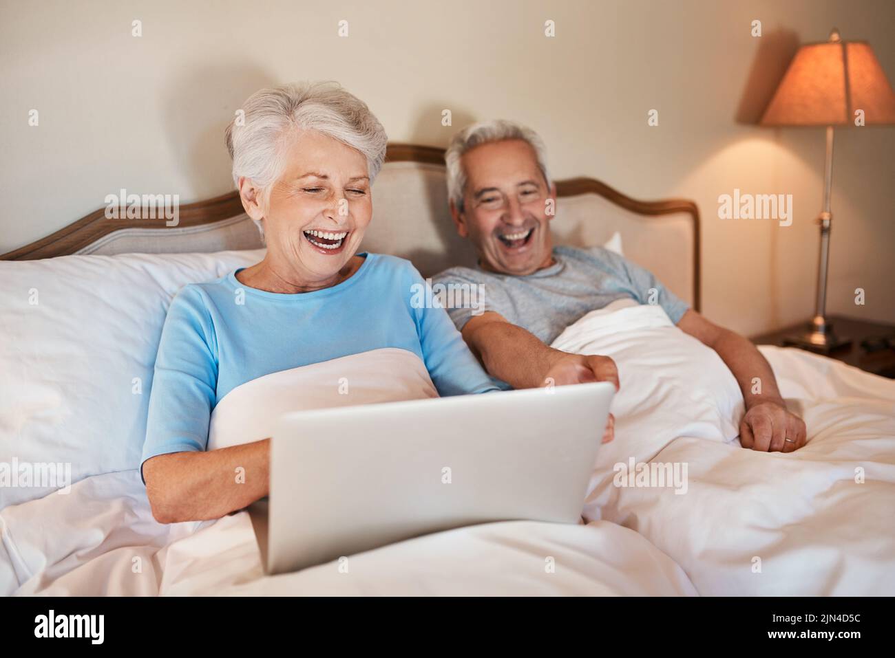 Oh, wie lustig die Enkel hier sind. Eine glückliche ältere Frau, die einen Laptop benutzt und zu Hause mit ihrem Mann im Bett sitzt. Stockfoto