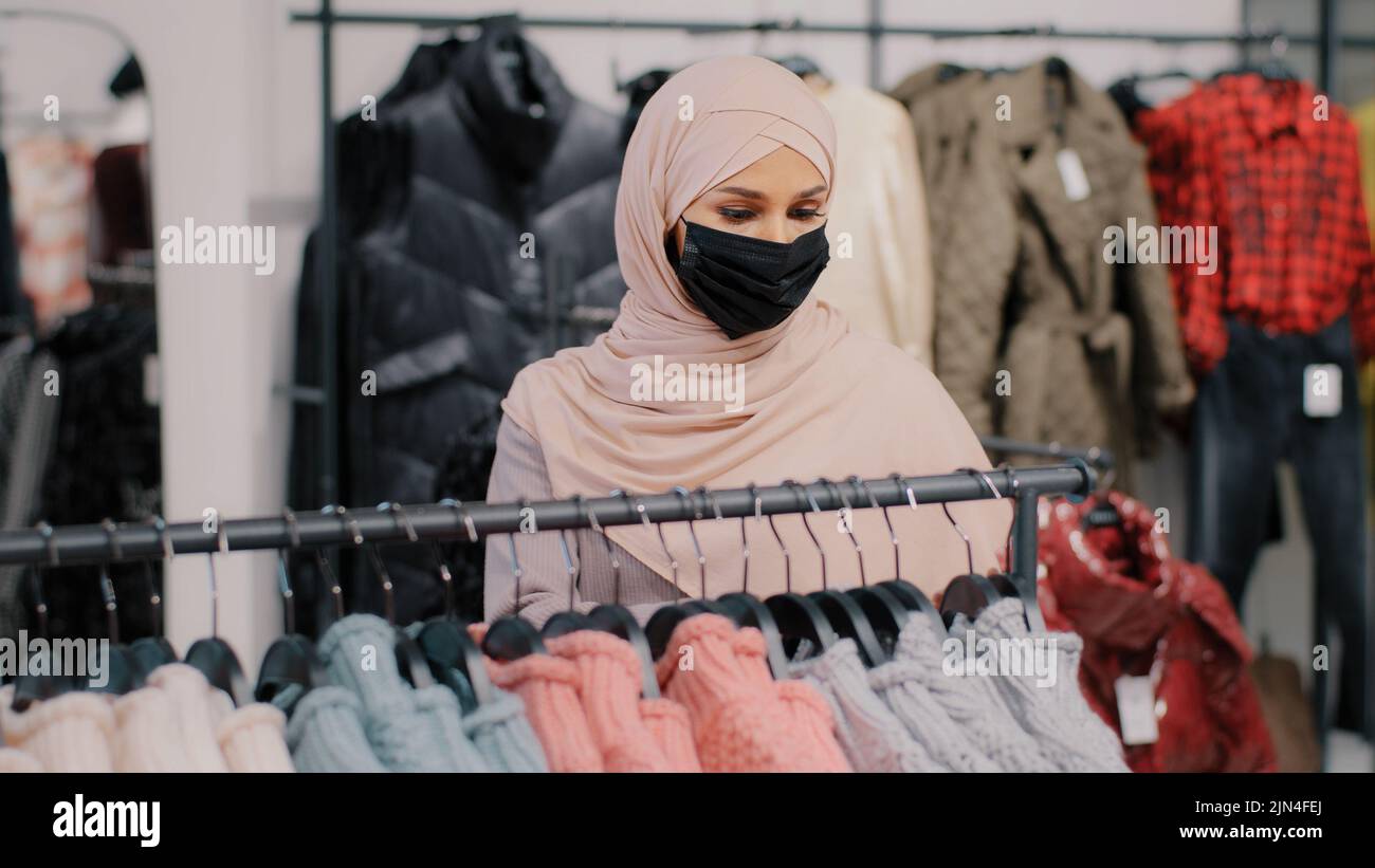 Junge arabische Frau Shopper in medizinische Schutzmaske wählt Kleidung in Bekleidungsgeschäft Einzelhandel Rabatt Kauf Einkaufen in Boutique muslim Stockfoto