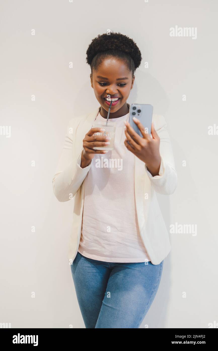 Lächelnde afro-amerikanische Frau, die einen Videoanruf auf dem Handy macht, ein Glas Getränk trinkt, isoliert auf weißem Hintergrund. Kreative neue Idee und Stockfoto