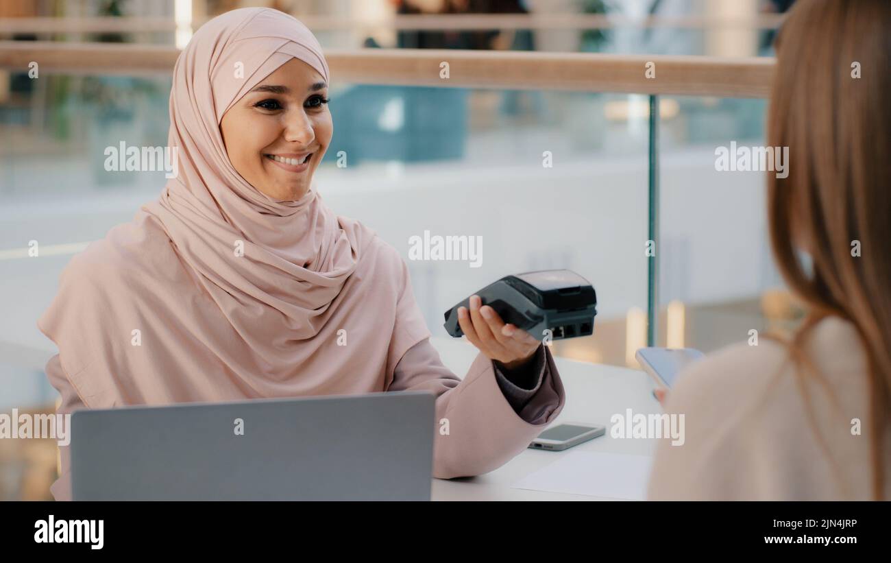 Freundliche muslimische Frau in Hijab Verkäuferagent bietet Bezahlung für Service über Bank-Terminal Mädchen Käufer Verbraucher zahlt für den Kauf mit kontaktlos Stockfoto